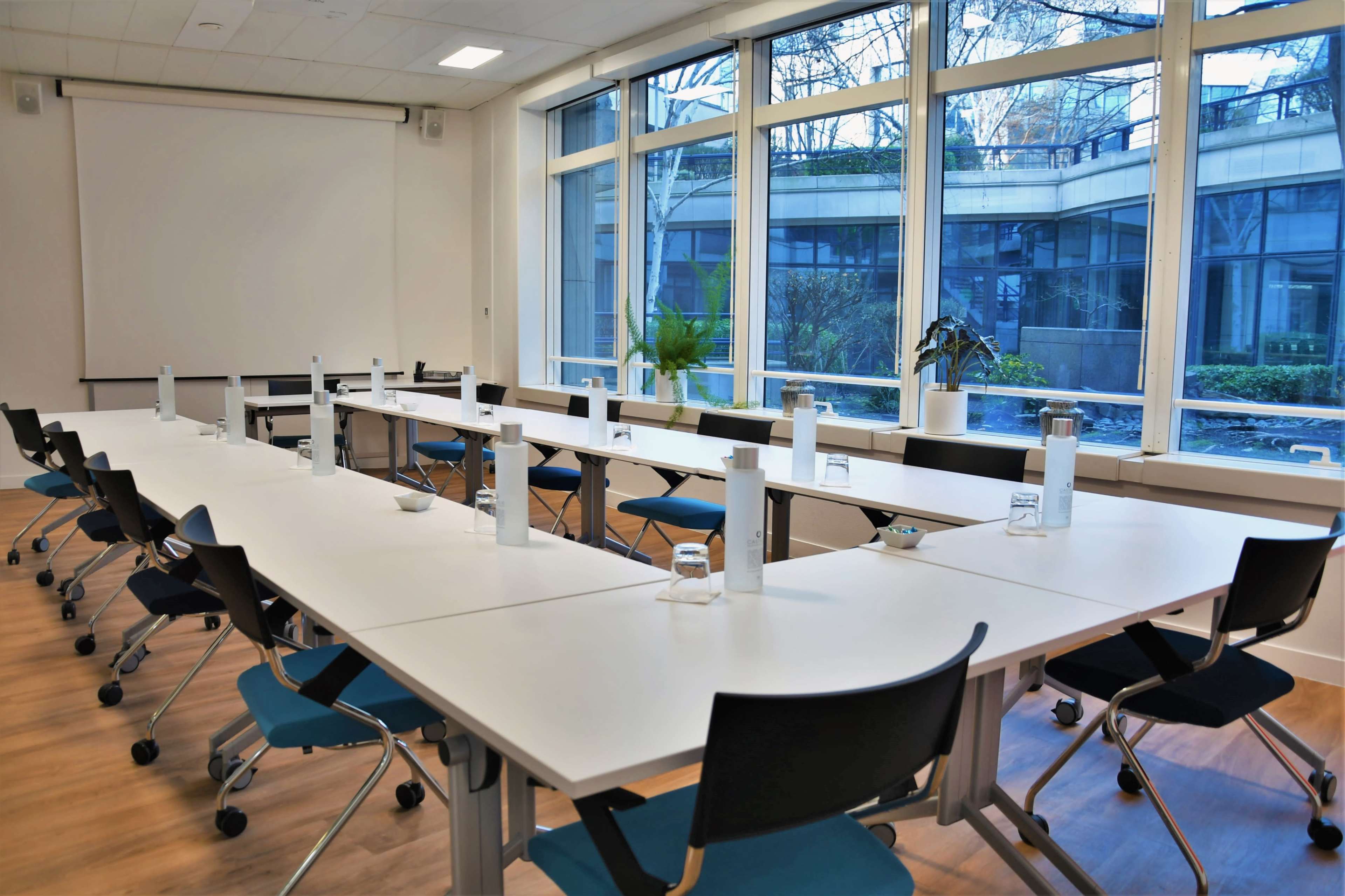 A large, modern conference room features a U-shaped arrangement of tables with chairs, glass windows, and plants in the background.