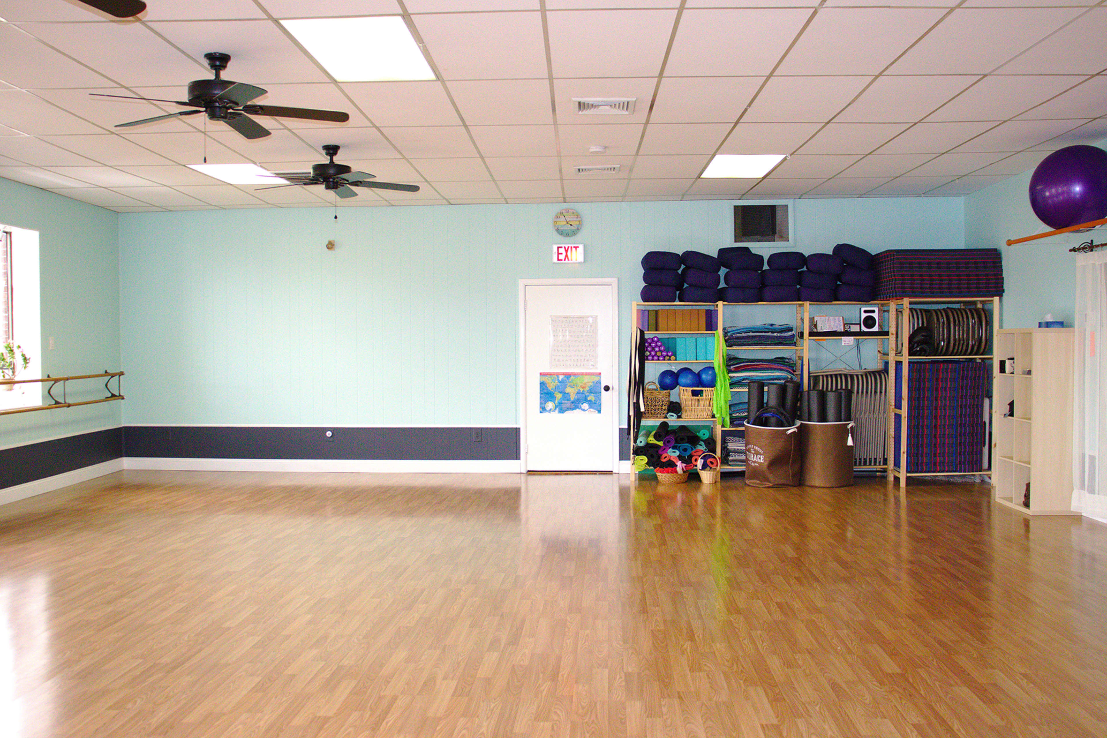 The image shows an empty fitness studio with wooden floors, a wall-mounted ballet bar, and shelves stacked with exercise equipment and mats.