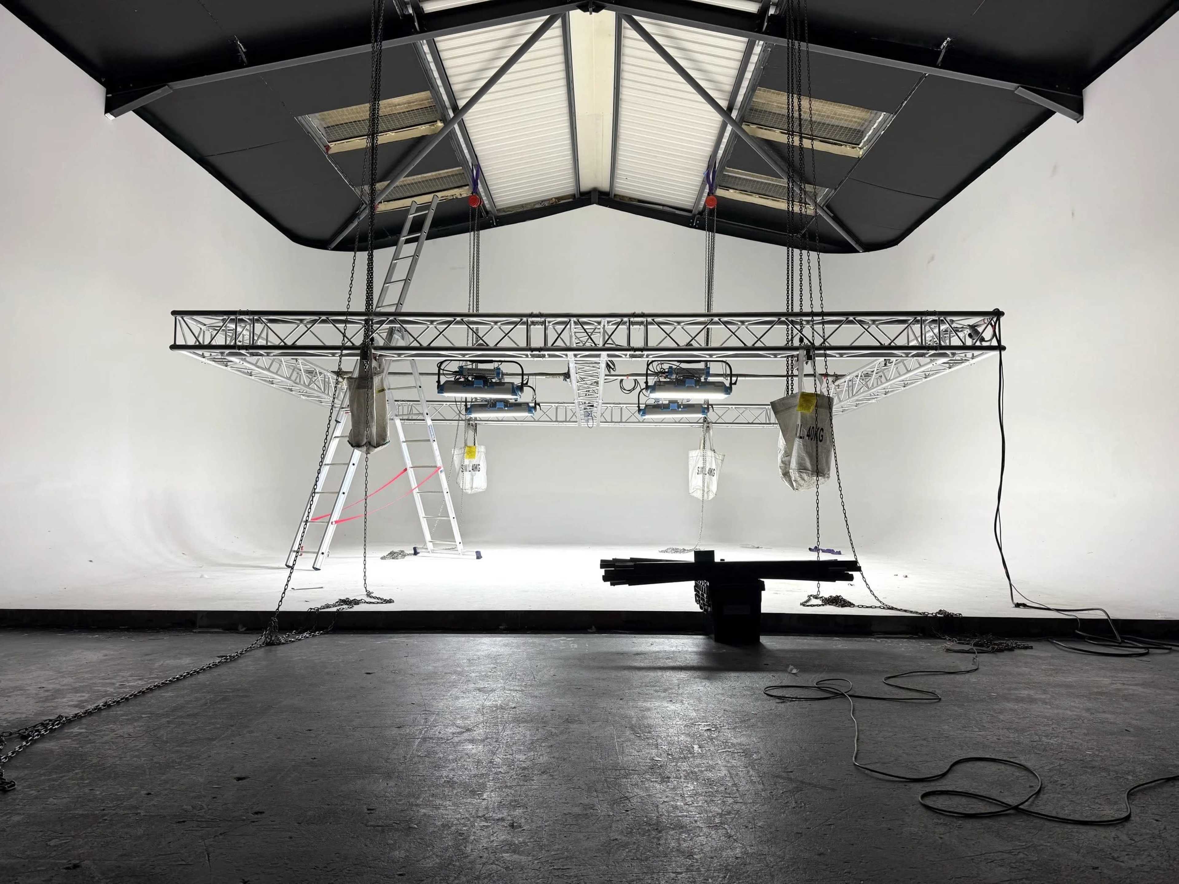 A studio space features a metal truss structure suspended from the ceiling with lighting fixtures and ladders positioned nearby.