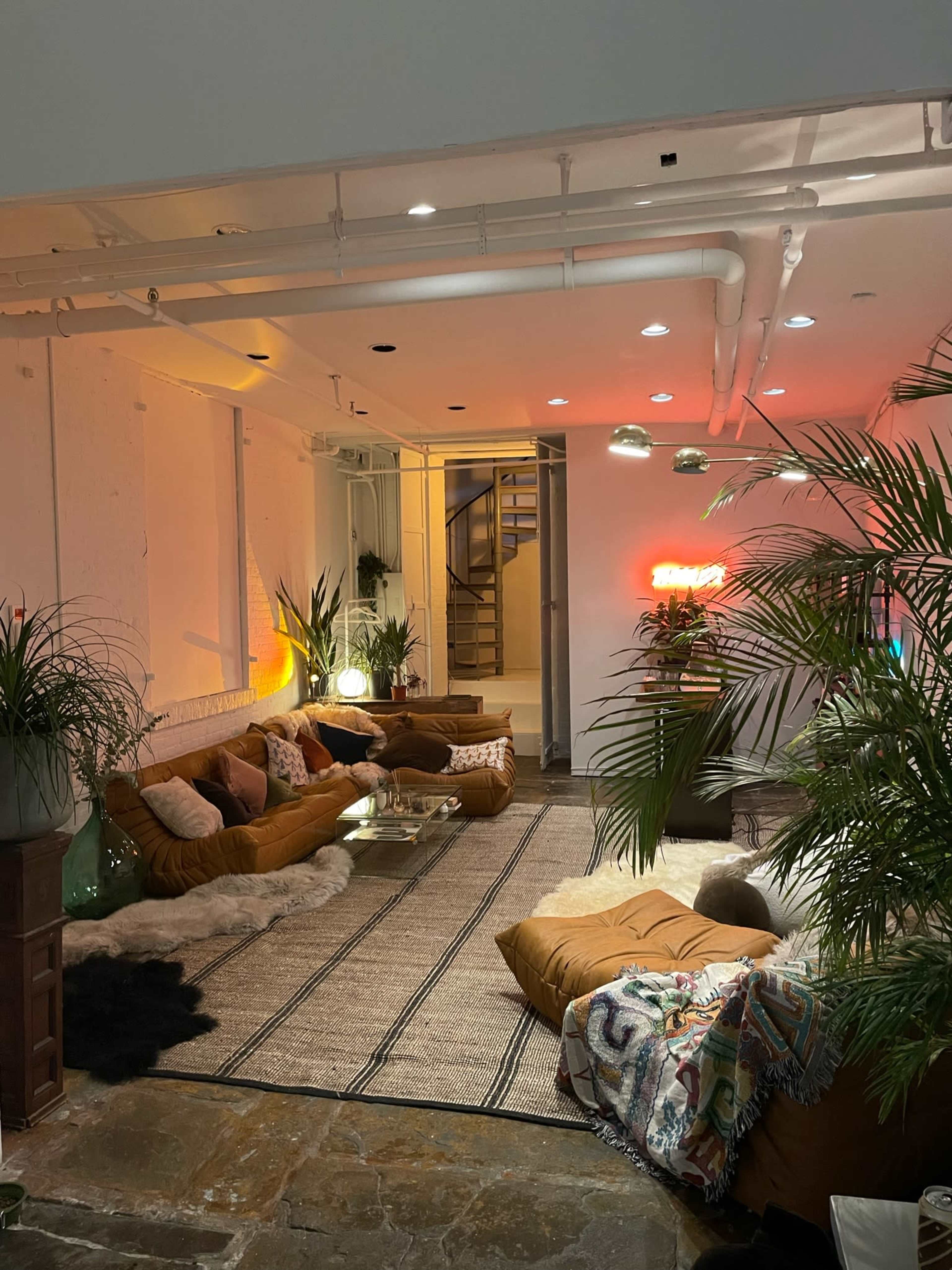 A cozy lounge area with large brown cushions, a glass coffee table, and indoor plants, leading to a staircase in the background.