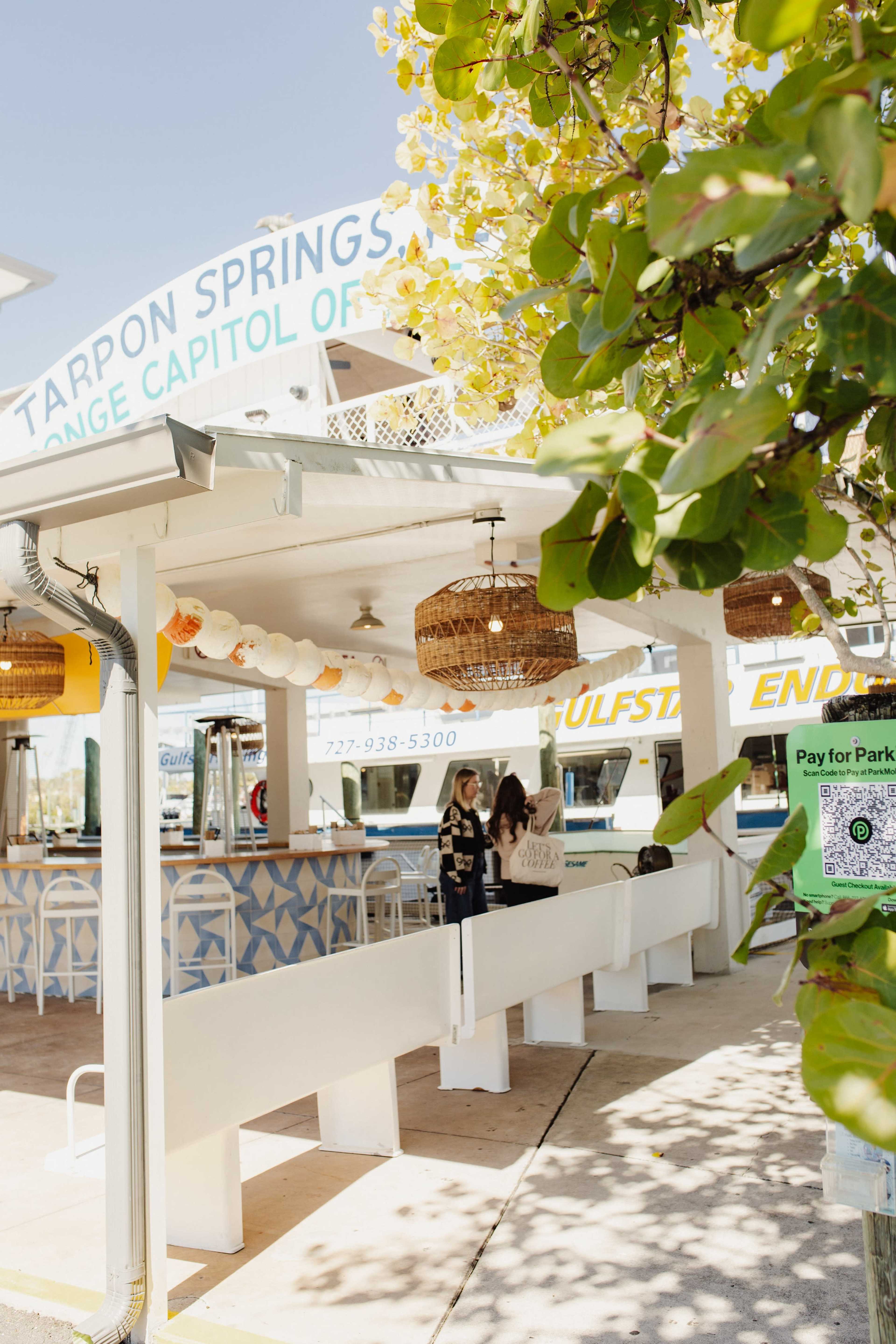 The image shows an outdoor dining area of a seaside restaurant with a wooden counter and decorative lighting, surrounded by greenery.