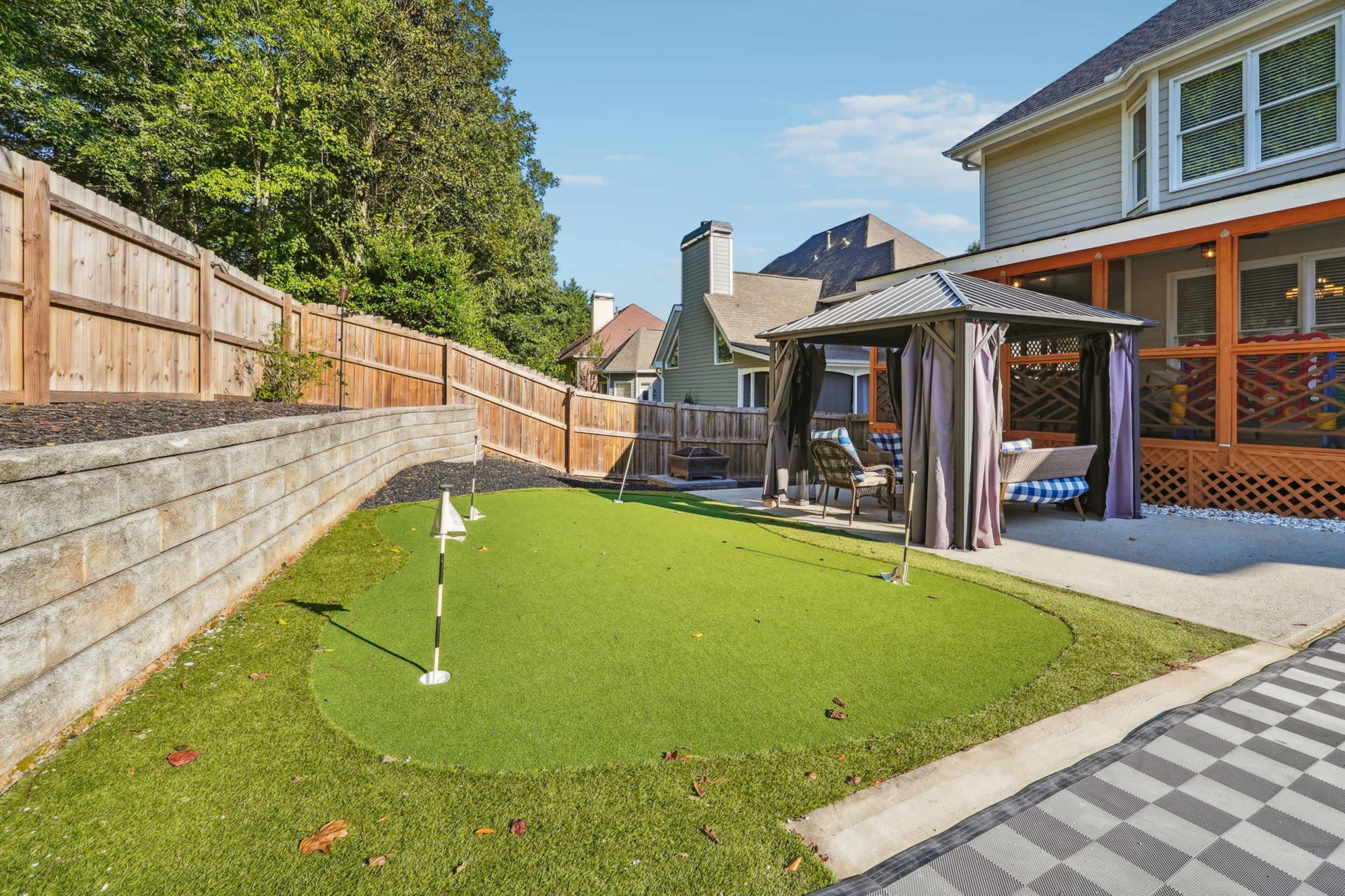 A backyard featuring a small artificial putting green with a white flag, a gazebo, and a stone wall surrounded by trees.