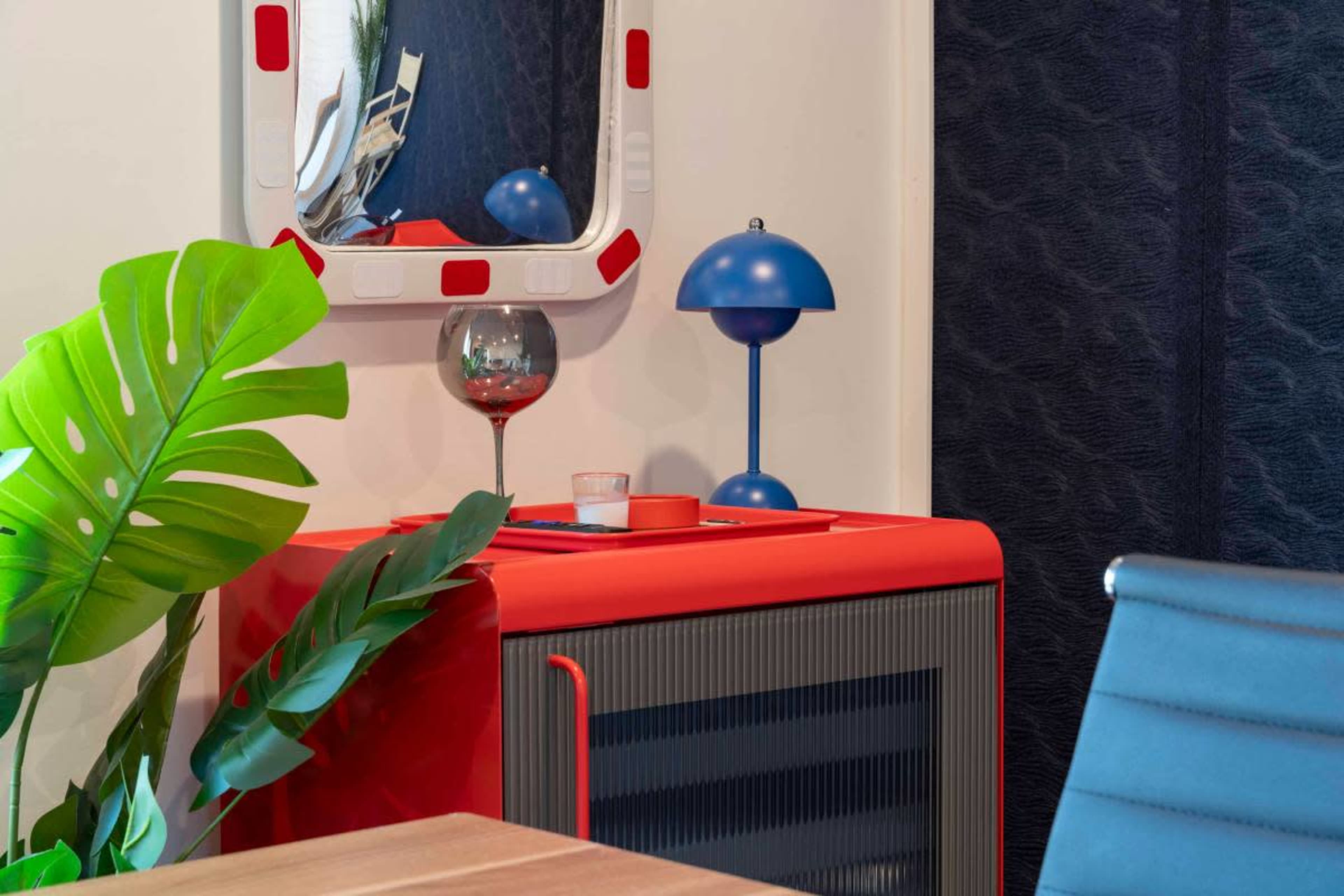 The image features a brightly colored red cabinet with a matching lamp, a glass of wine on top, and a large green plant, all set against a dark patterned wall.