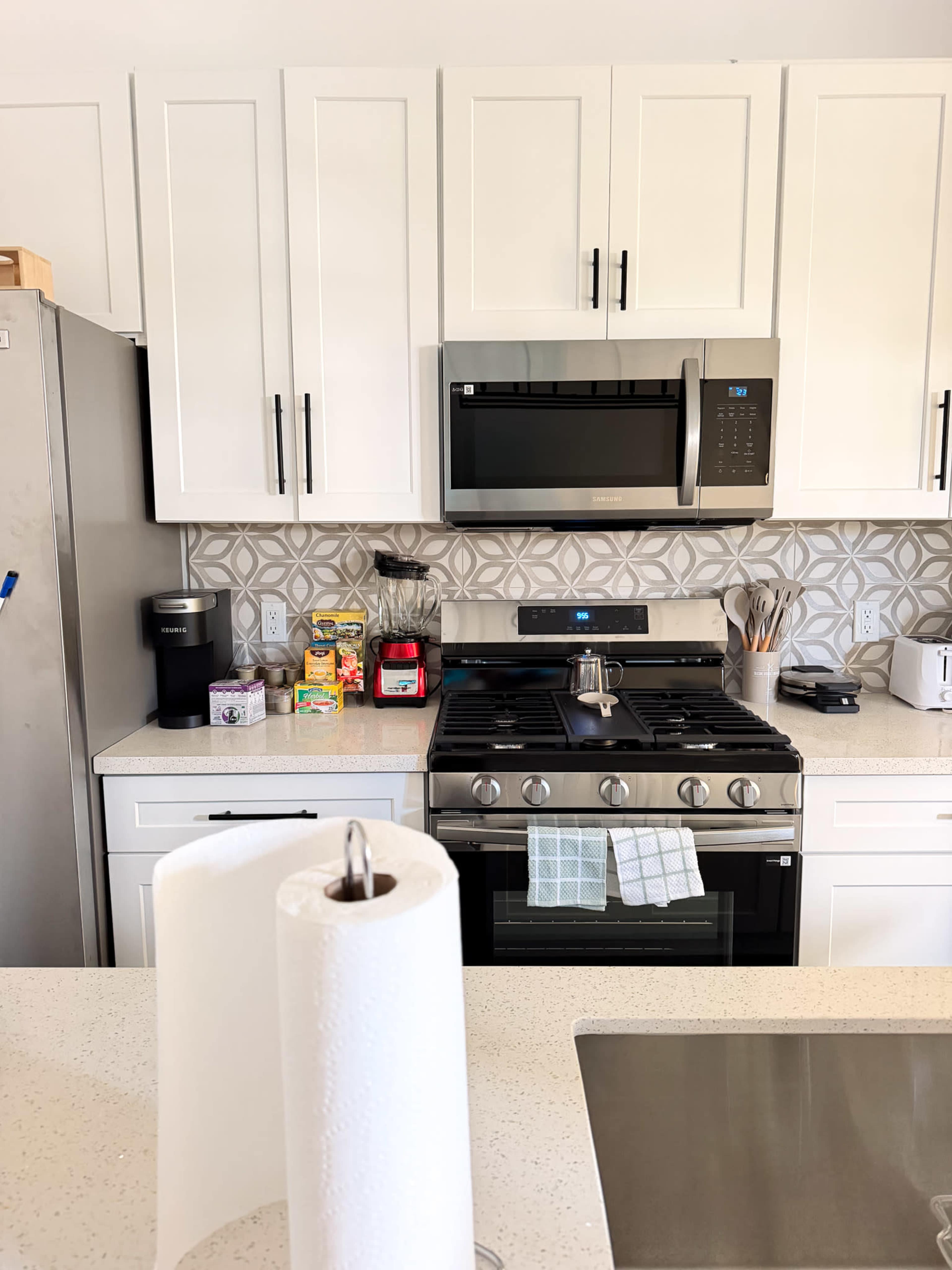 The image shows a modern kitchen with white cabinetry, stainless steel appliances, and a countertop featuring a roll of paper towels in the foreground.