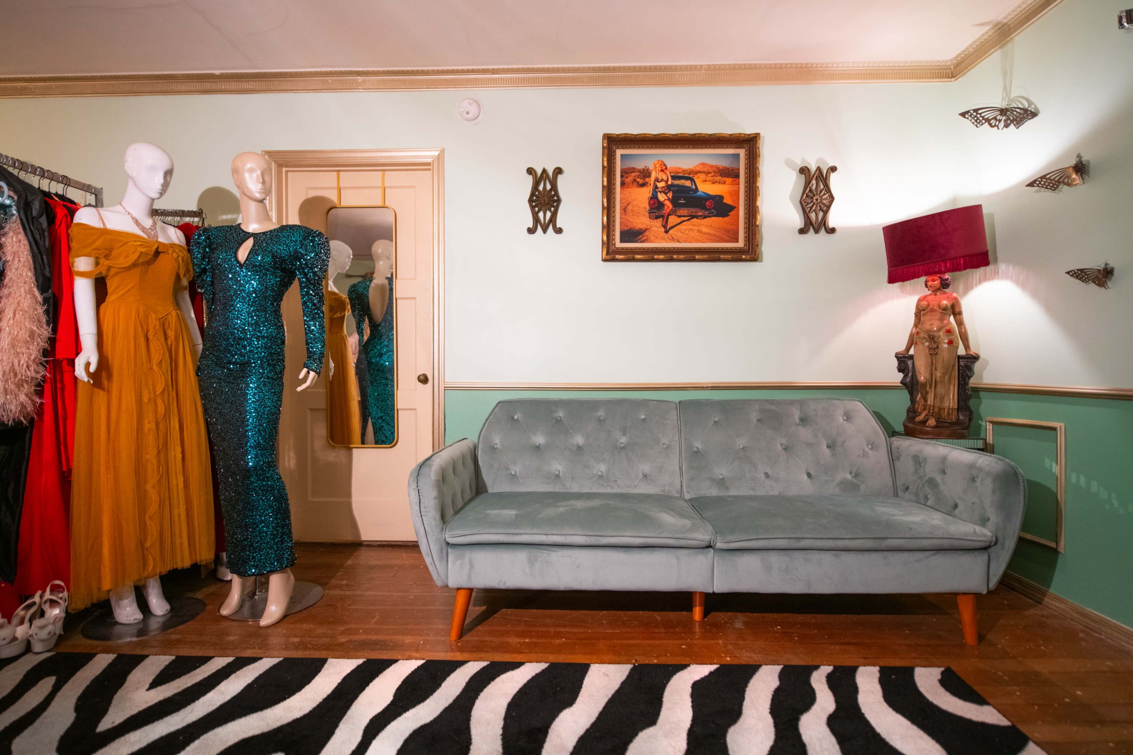 The scene features a vintage-inspired room with a green sofa, colorful dresses hanging on a rack, a decorative wall mirror, and a painting above an ornate lamp.