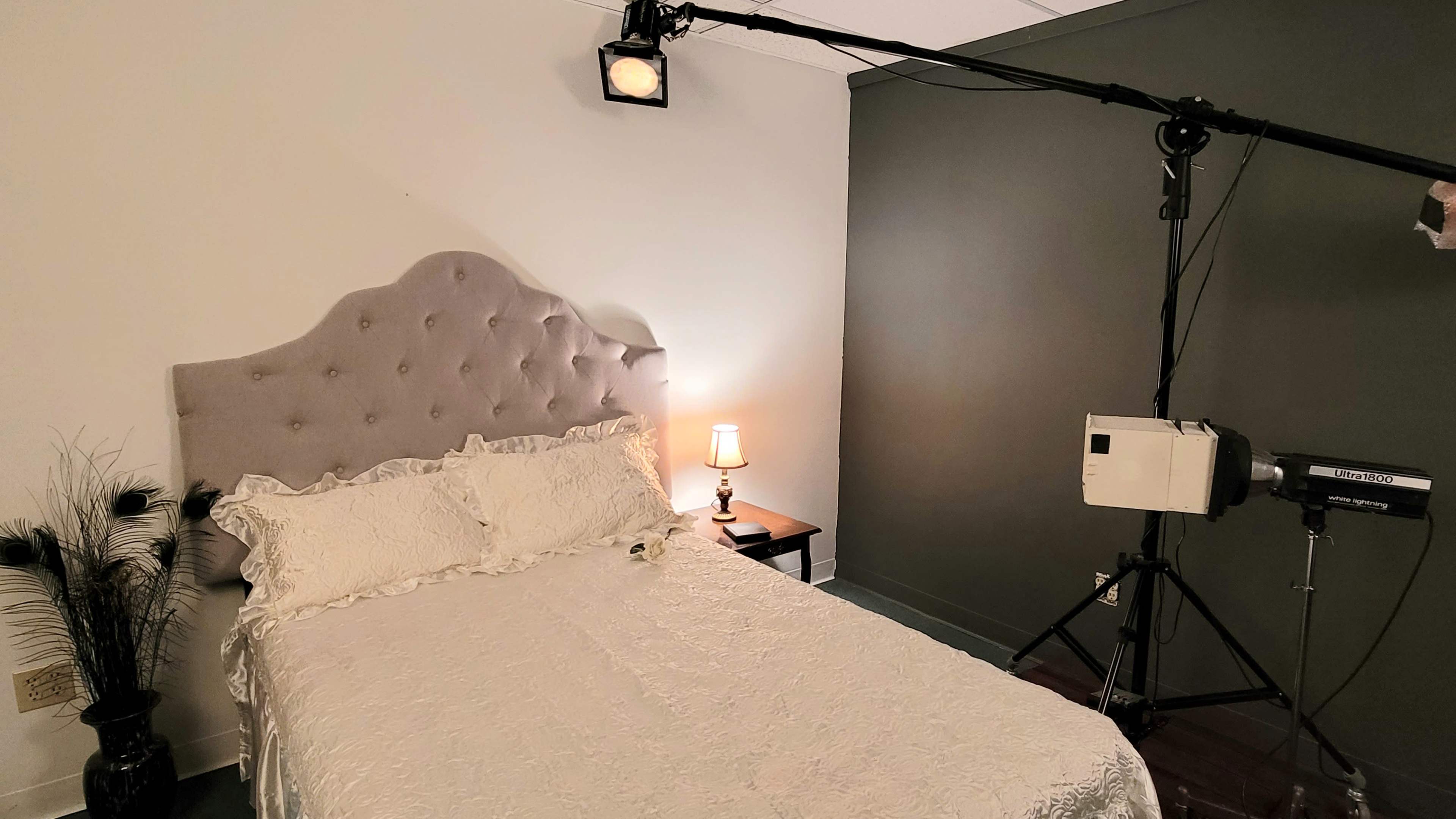 The image shows a neatly made bed with a gray headboard, a small nightstand with a lamp, and film equipment set up in a dimly lit room.