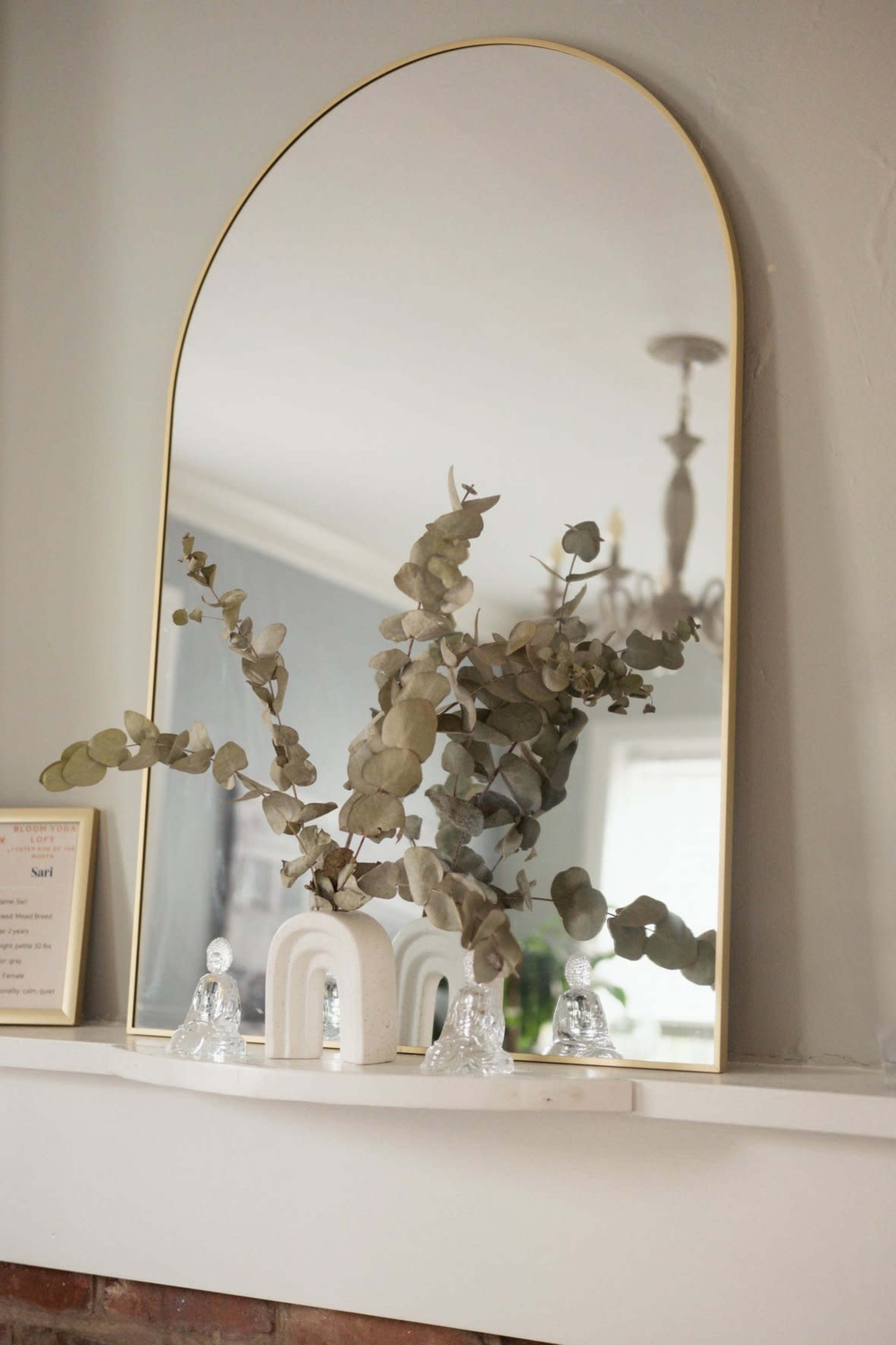 A large, arched mirror reflects a branch of eucalyptus and decorative glass figures on a mantelpiece.