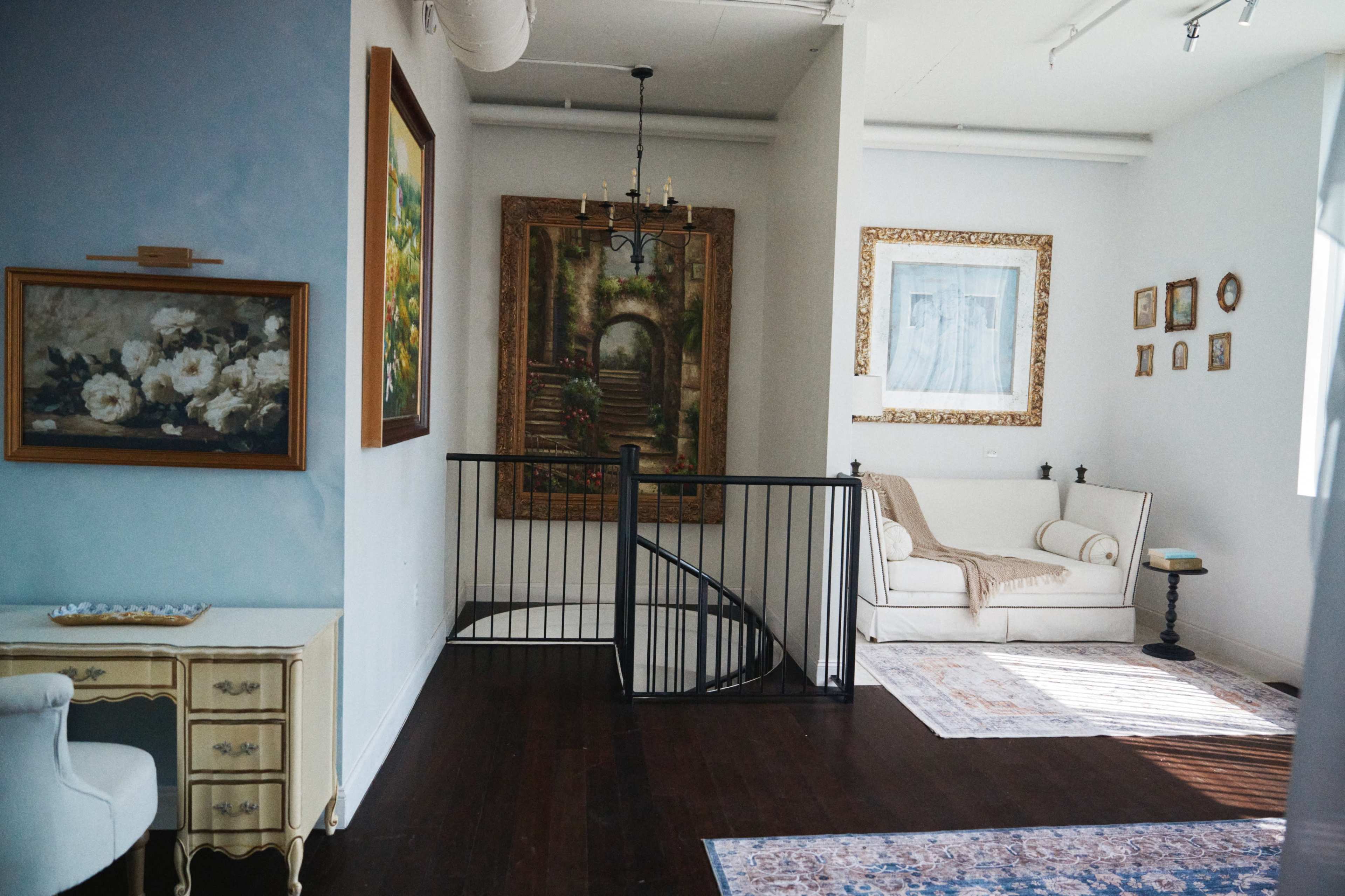 The image shows an interior space with a staircase, a white sofa, multiple framed artworks, and a small table in a well-lit room.
