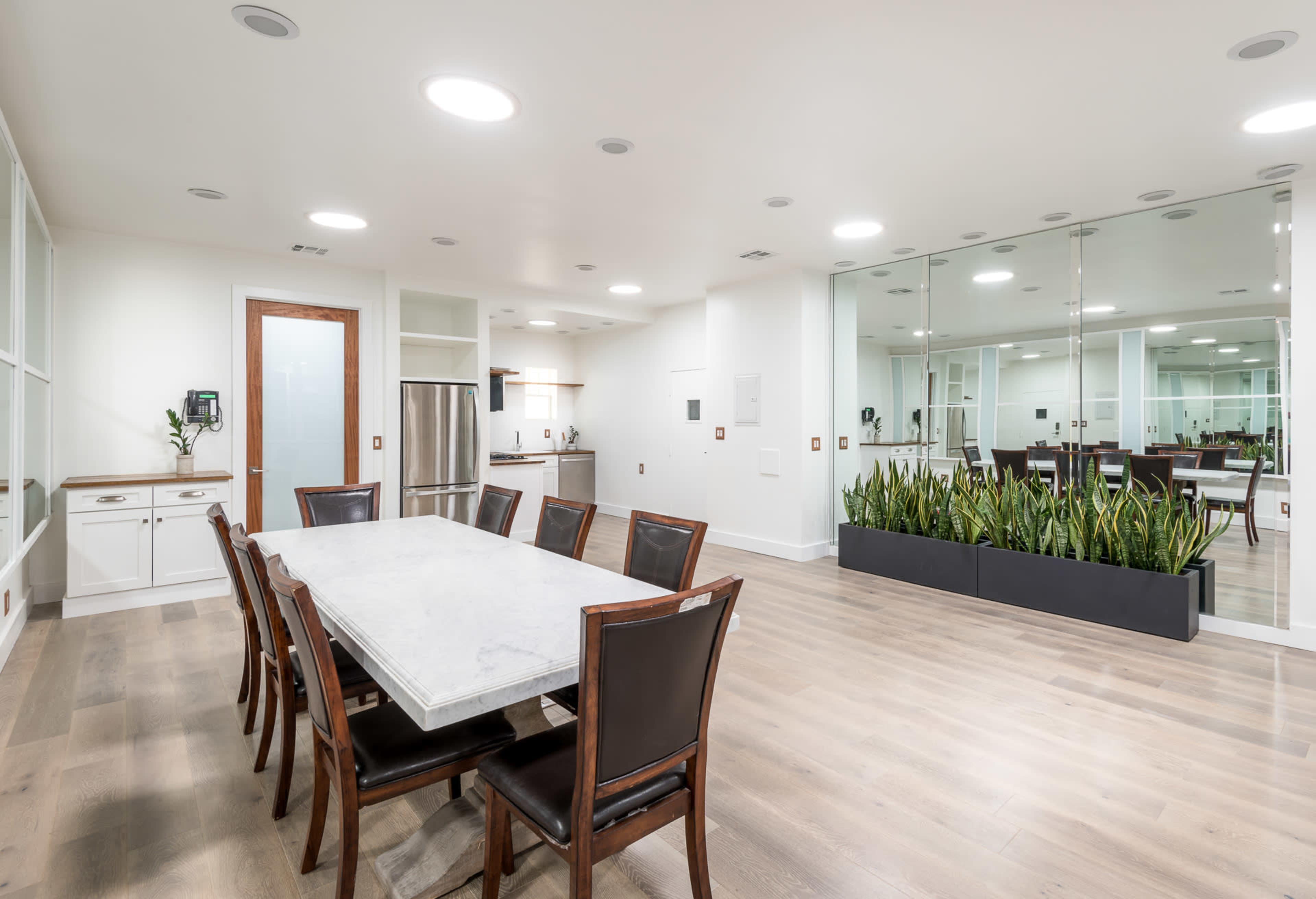 The image shows a modern dining area with a long marble table surrounded by eight chairs, adjacent to a kitchen space and a mirrored wall with decorative plants.