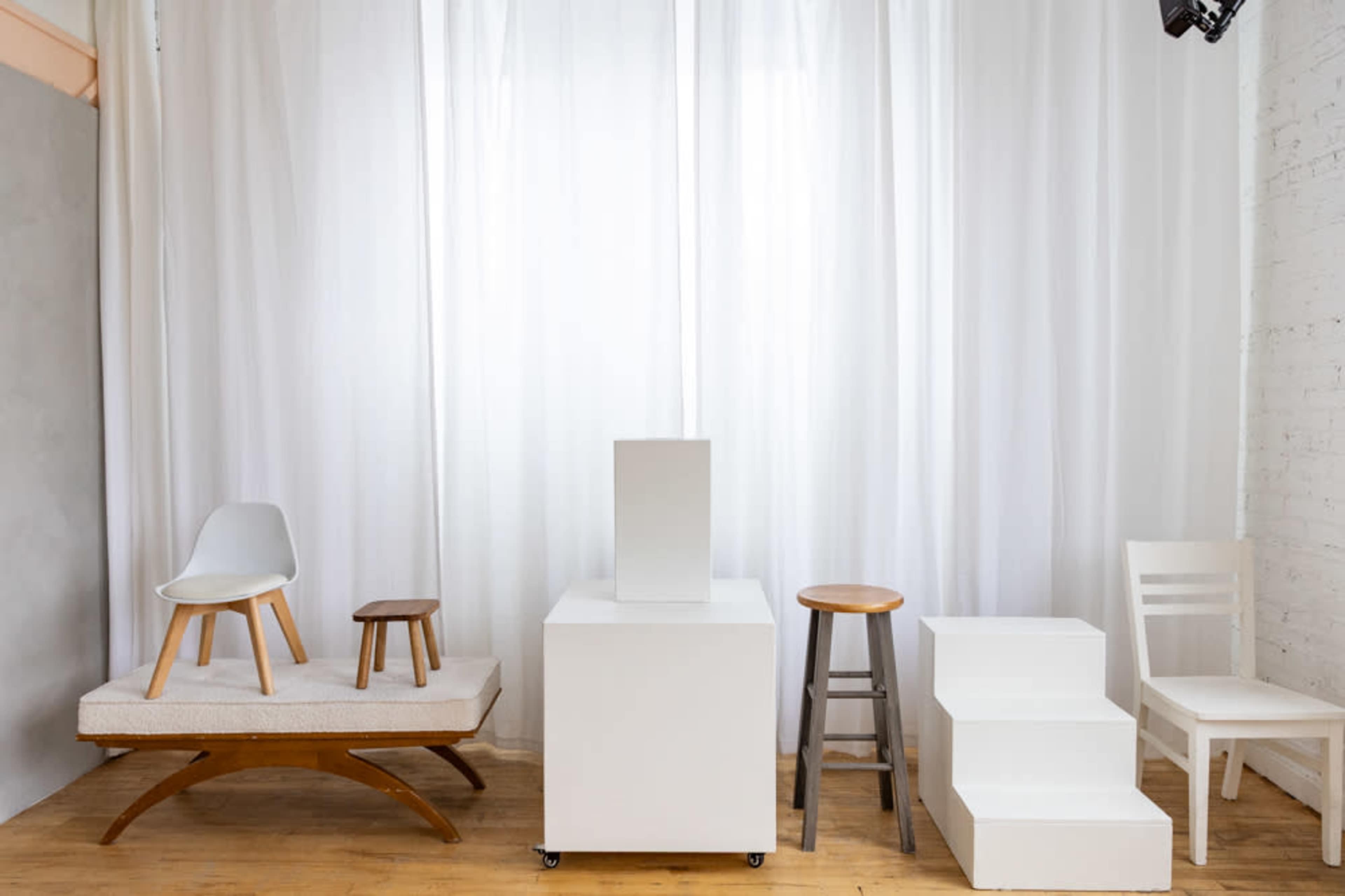 The image depicts a minimalist display featuring various types of furniture, including chairs and a stool, arranged in front of a white curtain backdrop.