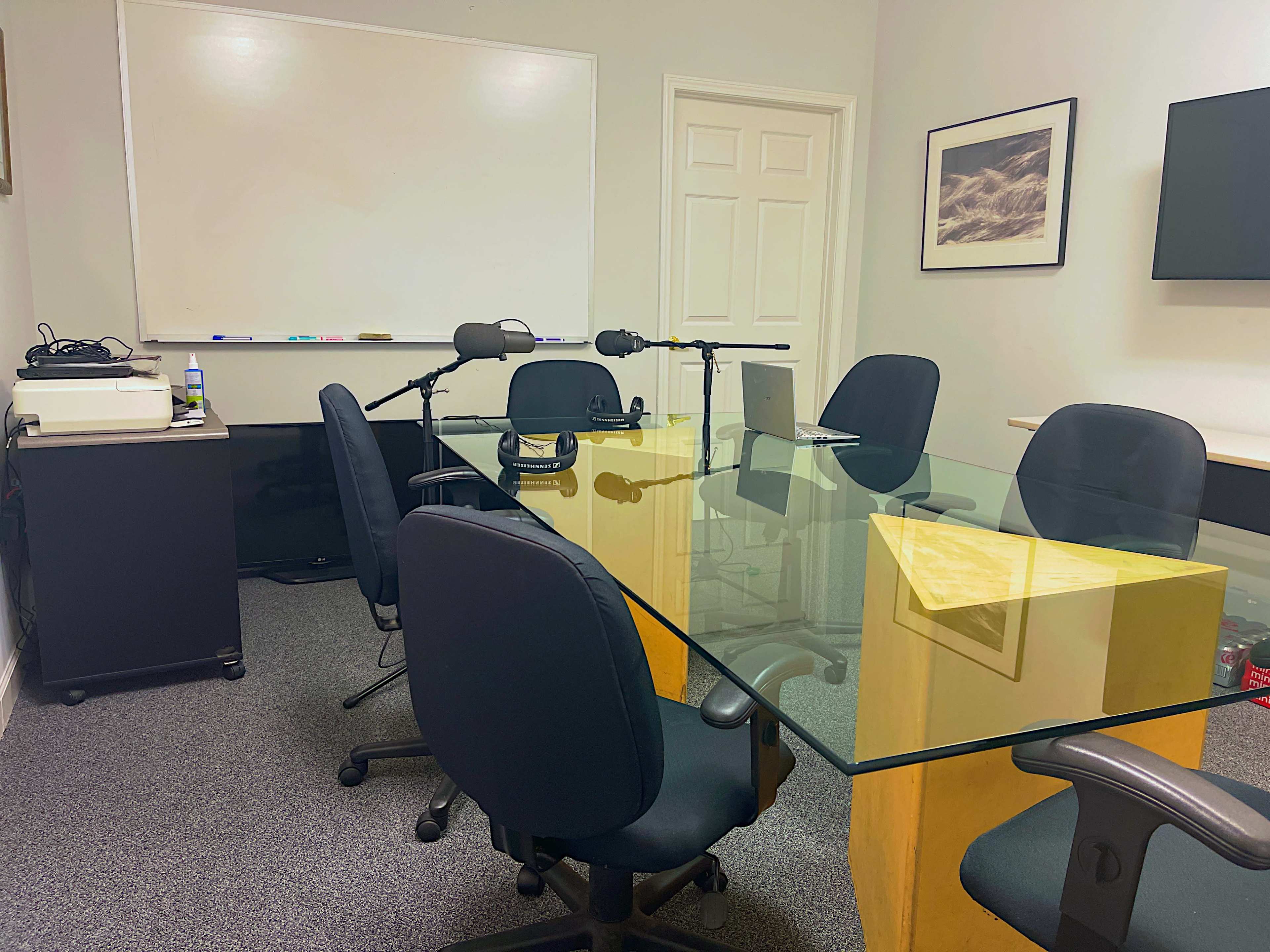 The image shows a meeting room with a glass table surrounded by black chairs, a whiteboard, and recording equipment arranged in the corner.