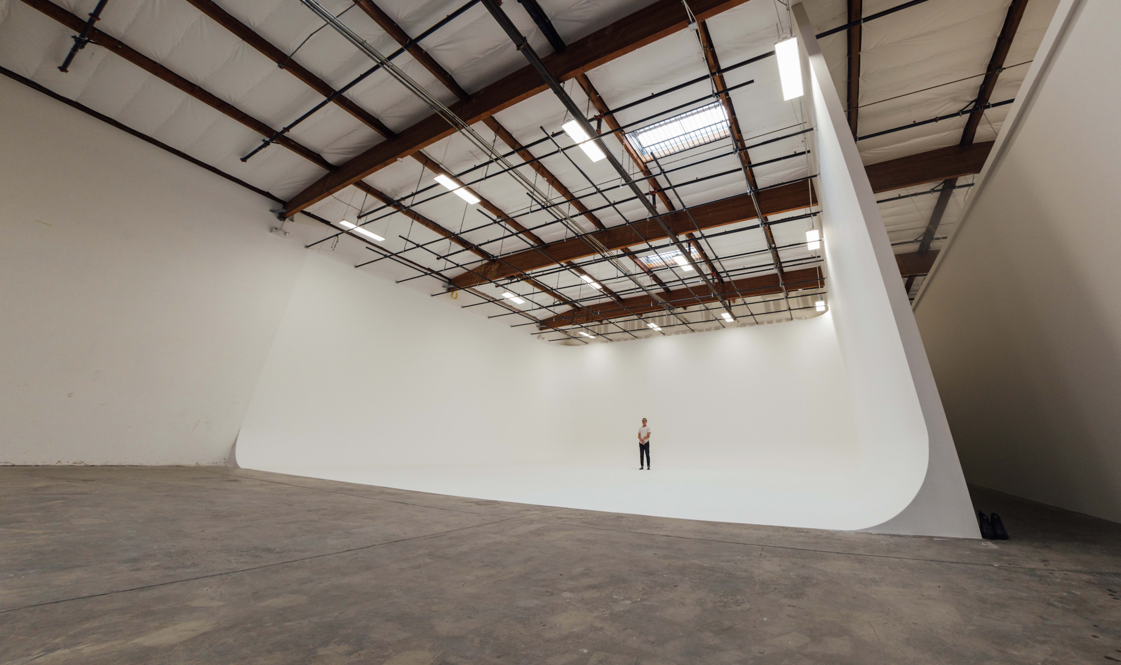 A person stands alone in a large, empty studio with a curved white backdrop and exposed wooden beams overhead.