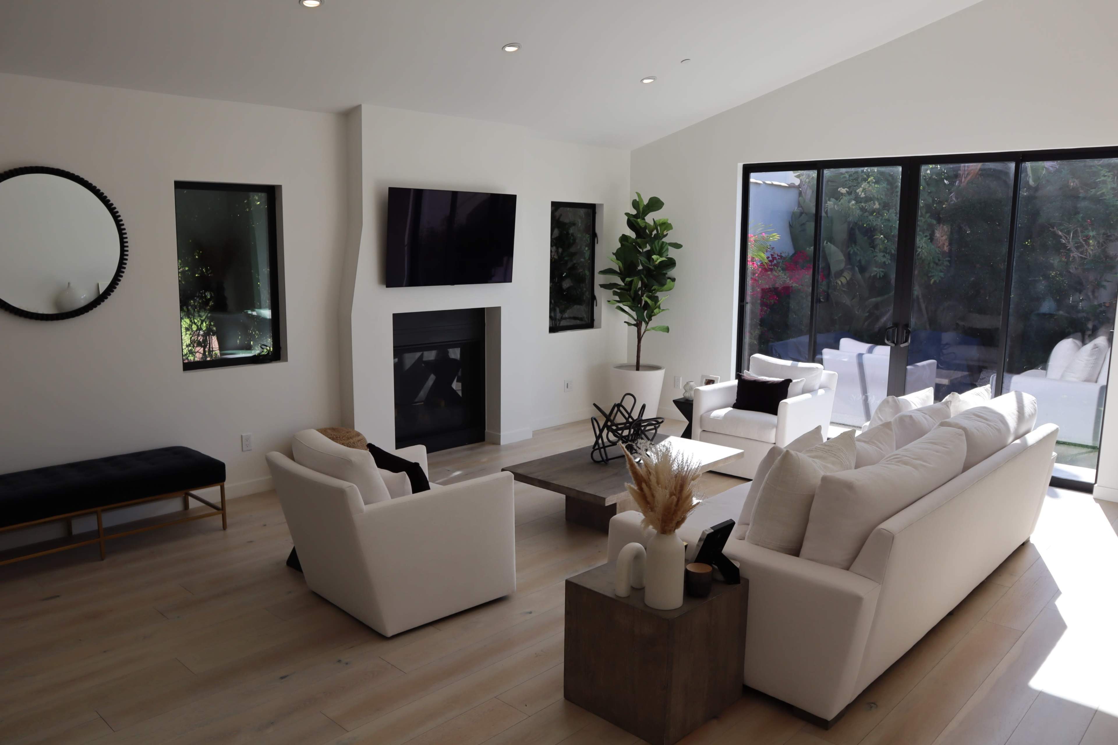 A modern living room features white furniture, a fireplace, a large window with garden views, and minimalistic decor.