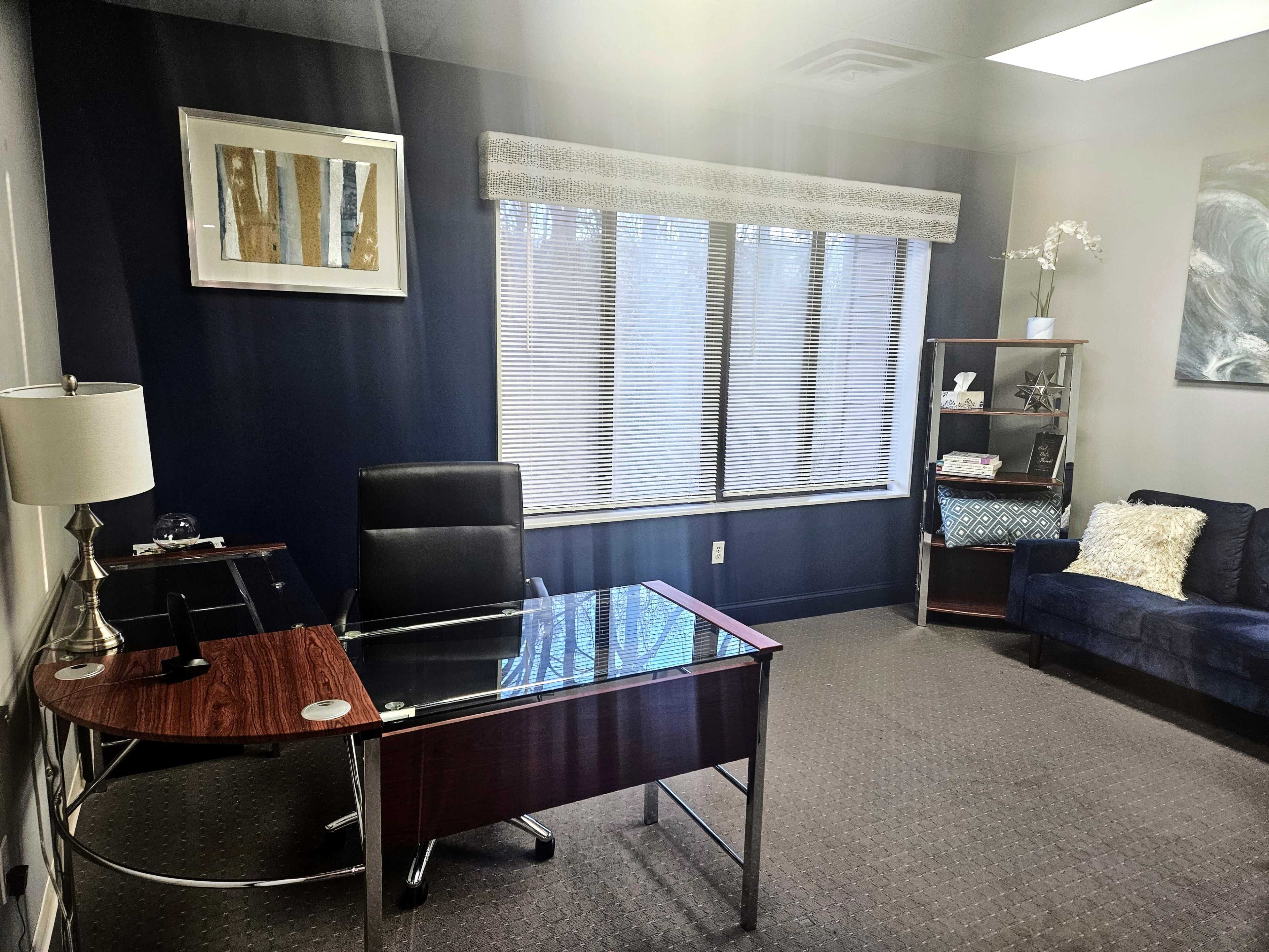 A minimalist office space featuring a glass-top desk, a black chair, a blue couch, and a window with blinds.