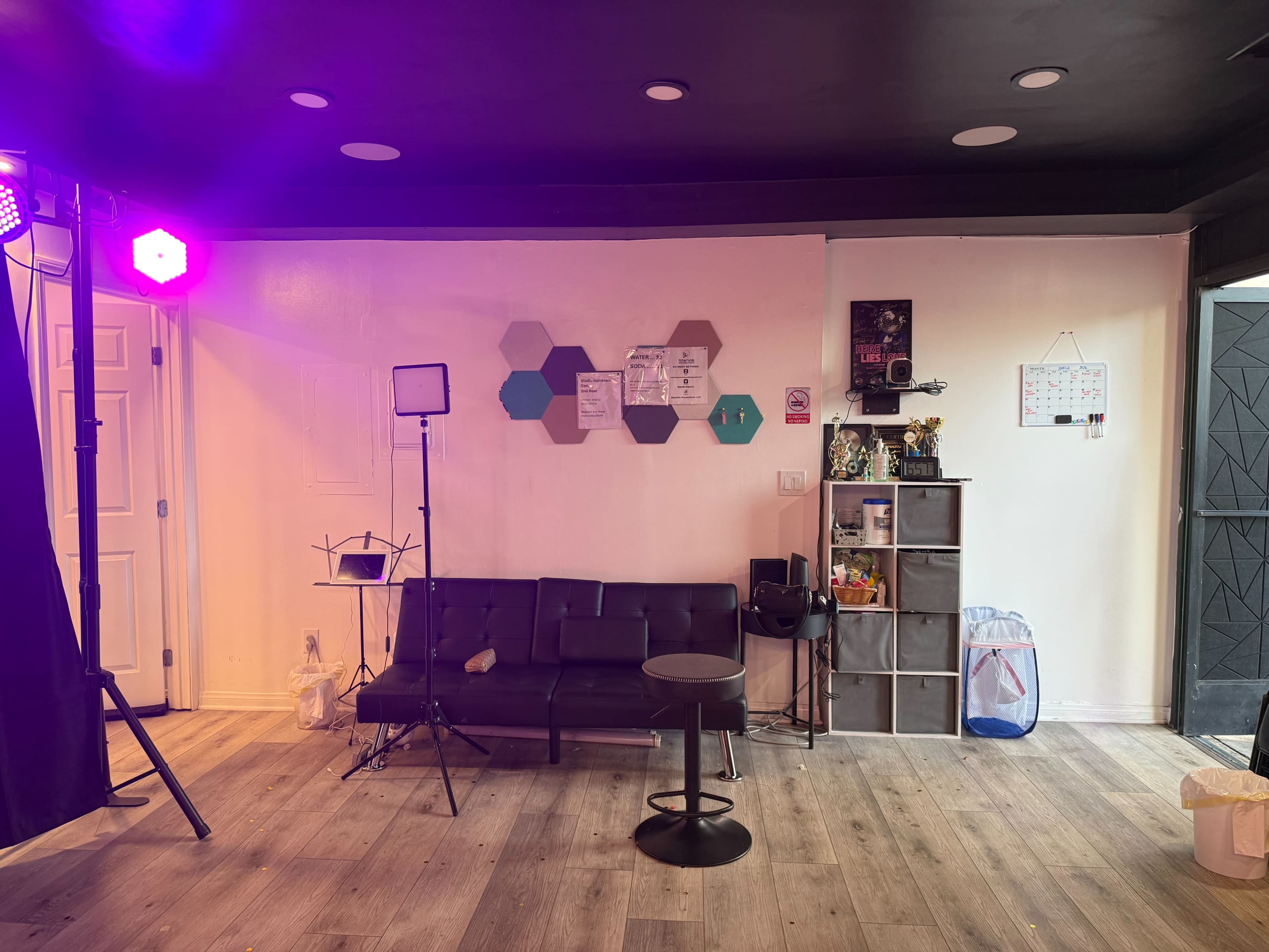 A modern interior space features a black couch, a small round table, a shelf with various items, and studio lighting set up against a light-colored wall.