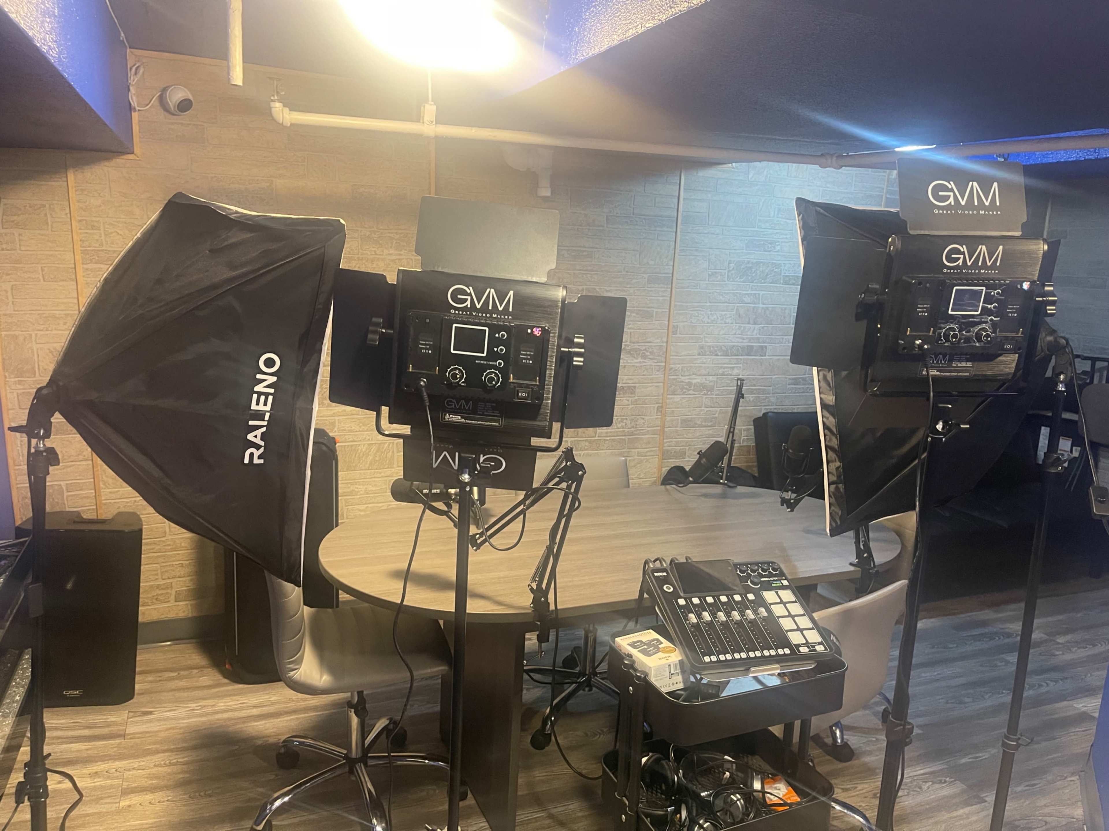 Two professional lighting setups with softboxes are positioned around a central table in a studio-like environment, accompanied by a control panel and chairs.