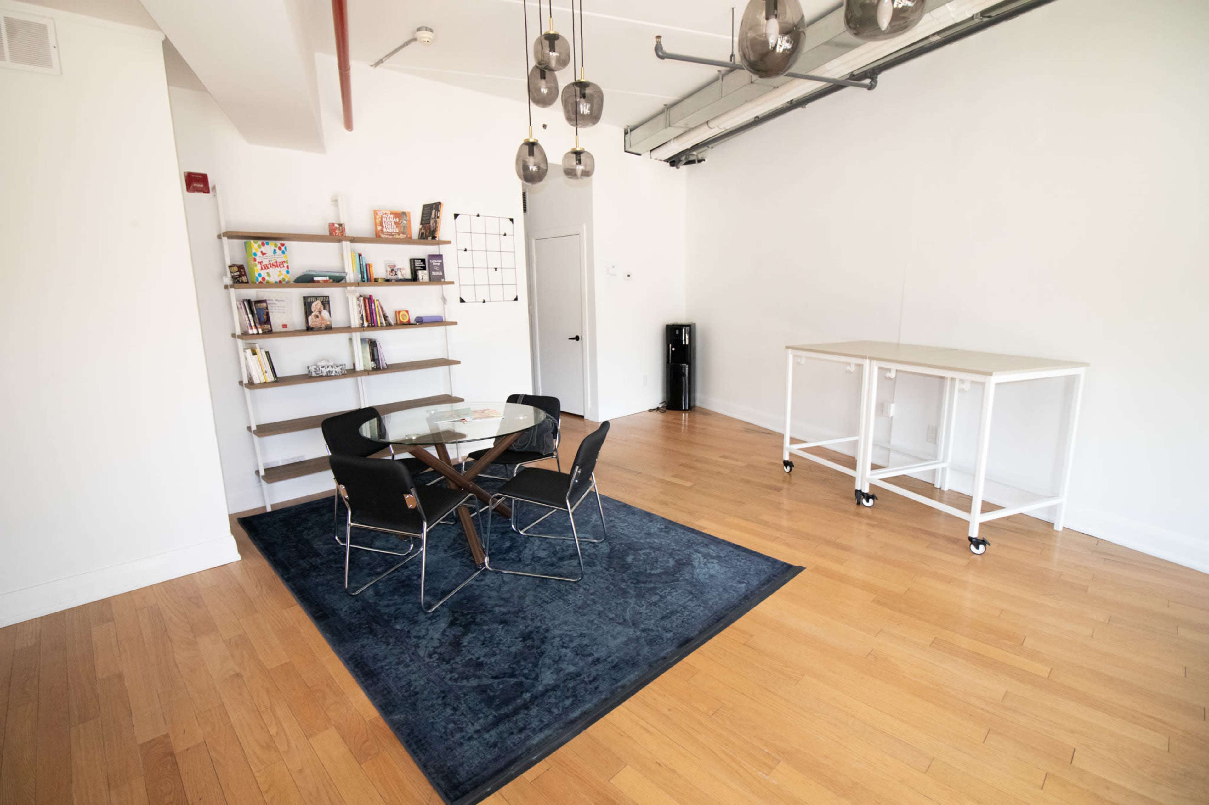 The image shows a modern, minimalistic interior featuring a wooden floor, a round table with four chairs, a bookshelf with various items, and a small white cart.