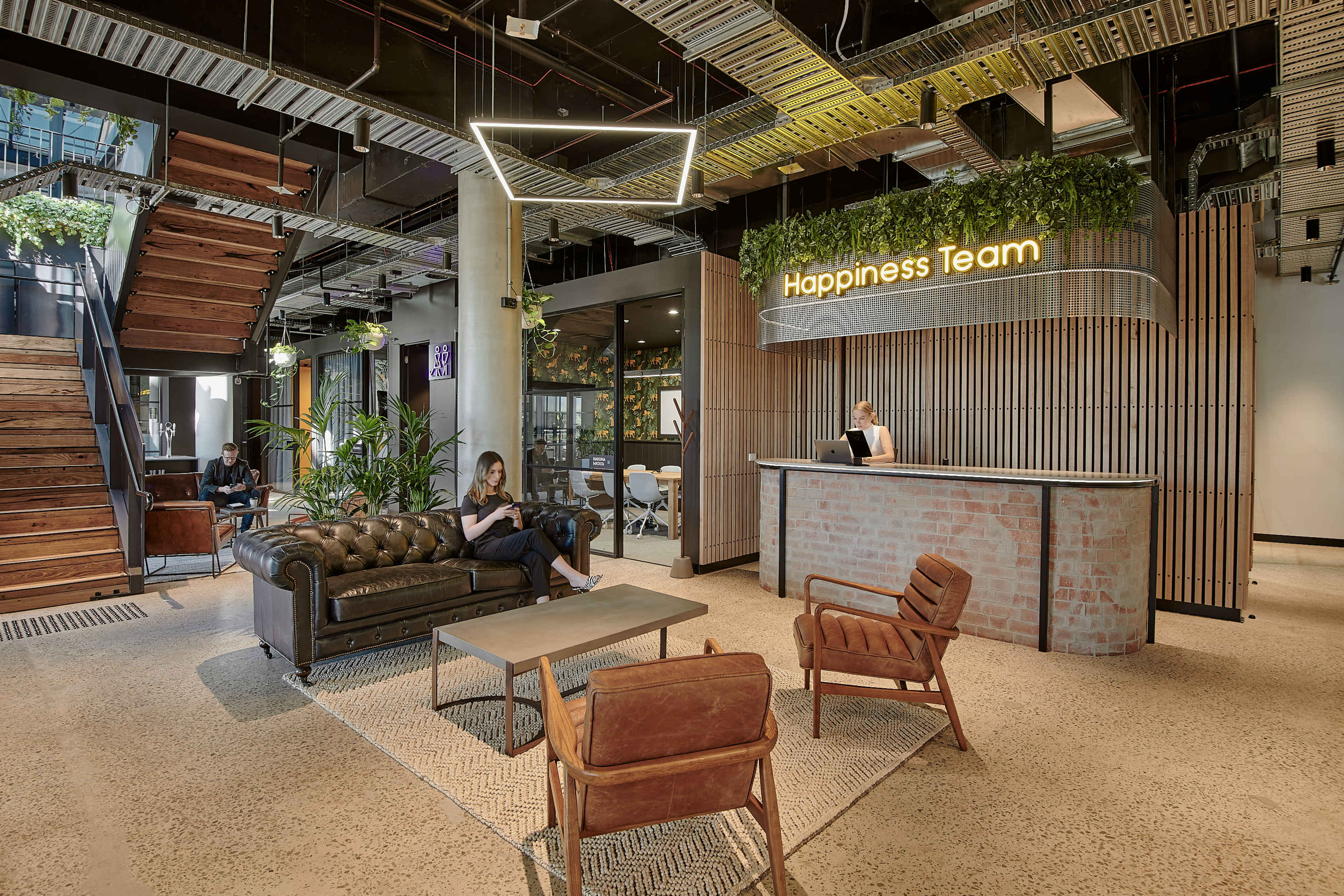 The image shows a modern office lobby with a reception desk, seating area, and a staircase, featuring plants and contemporary furniture.