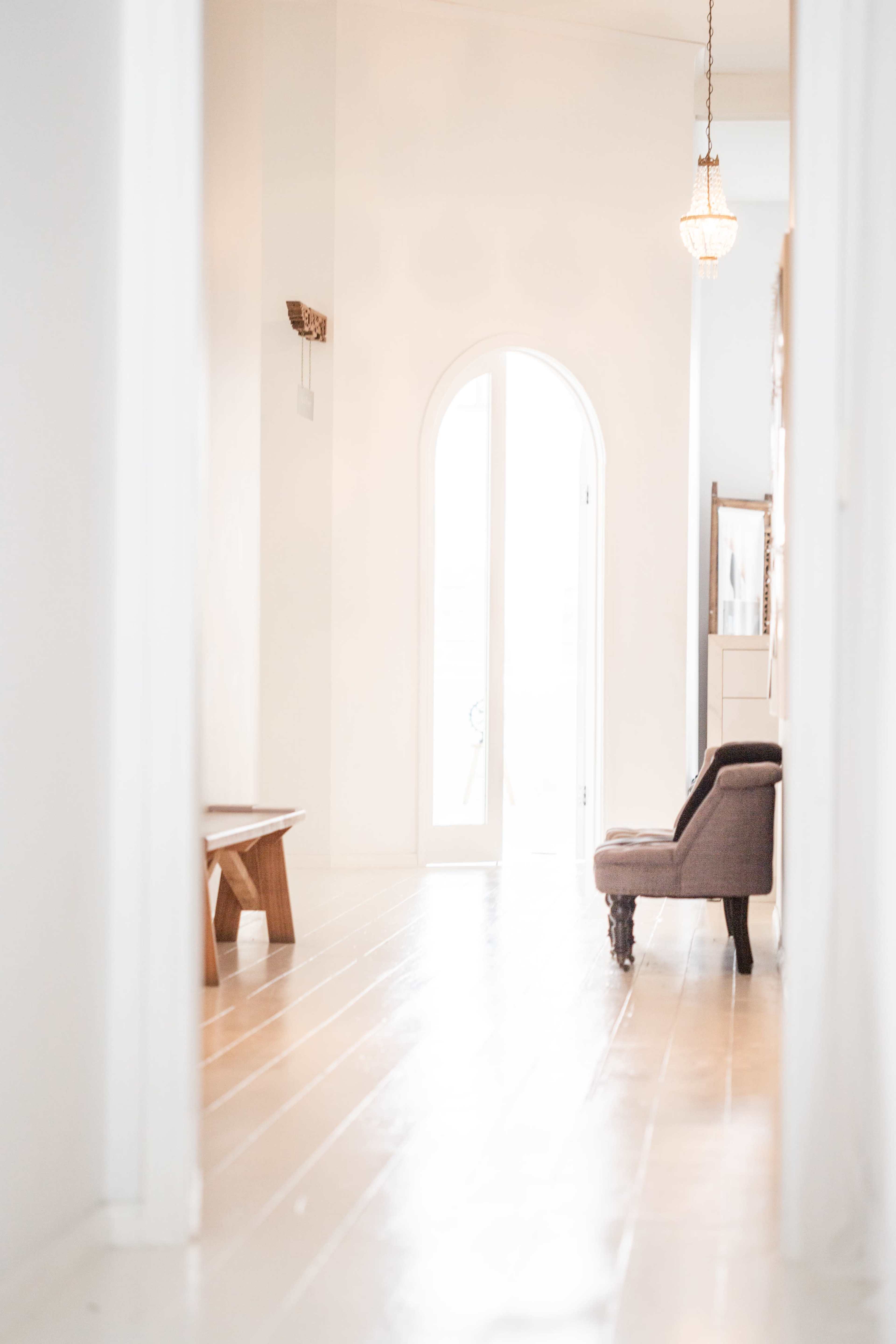 A narrow hallway leads to a light-filled archway, featuring a wooden bench and an upholstered chair along its walls.