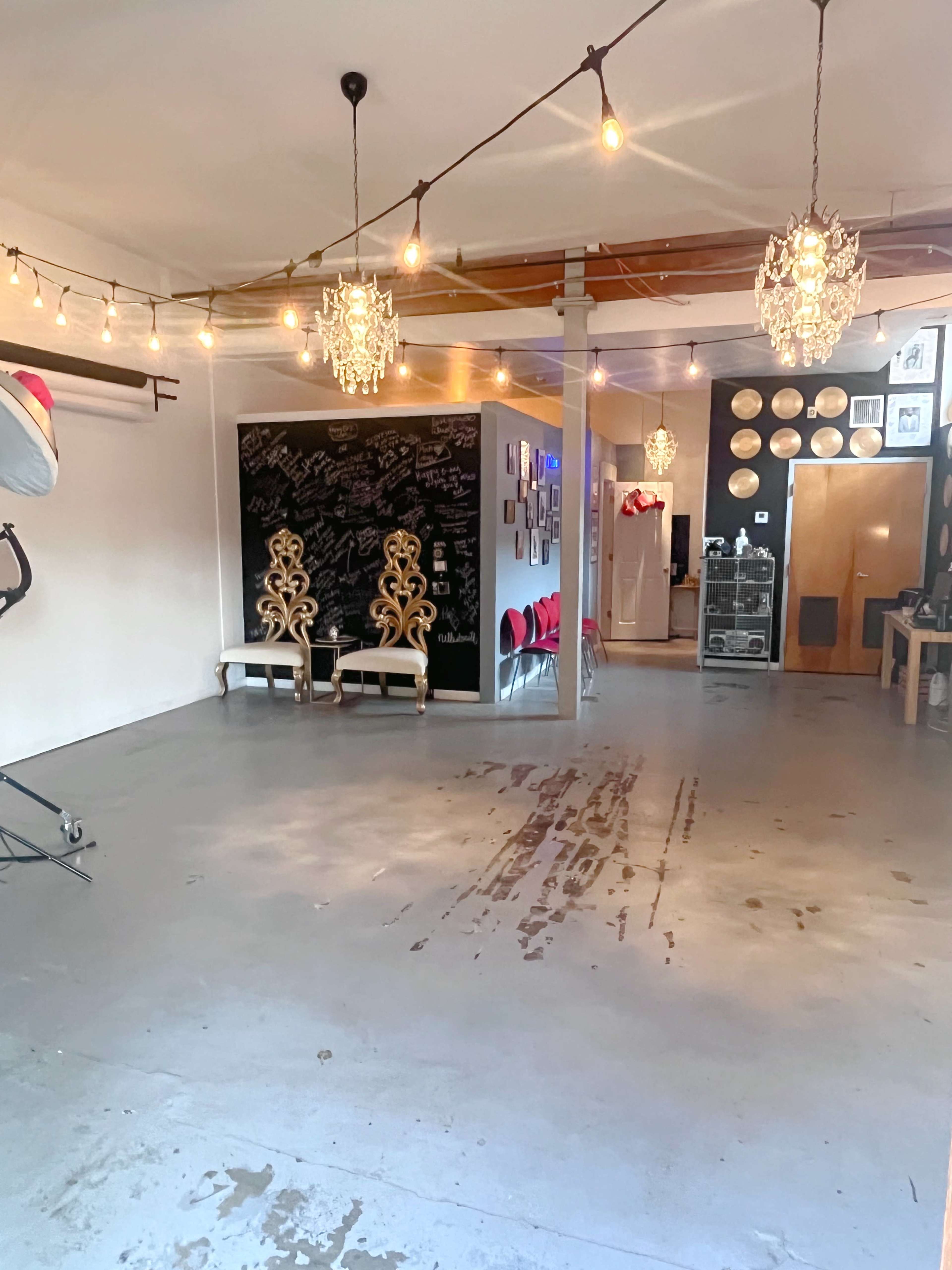 The image shows a spacious room with string lights, decorative chandeliers, a chalkboard wall, and various furniture pieces along the walls.