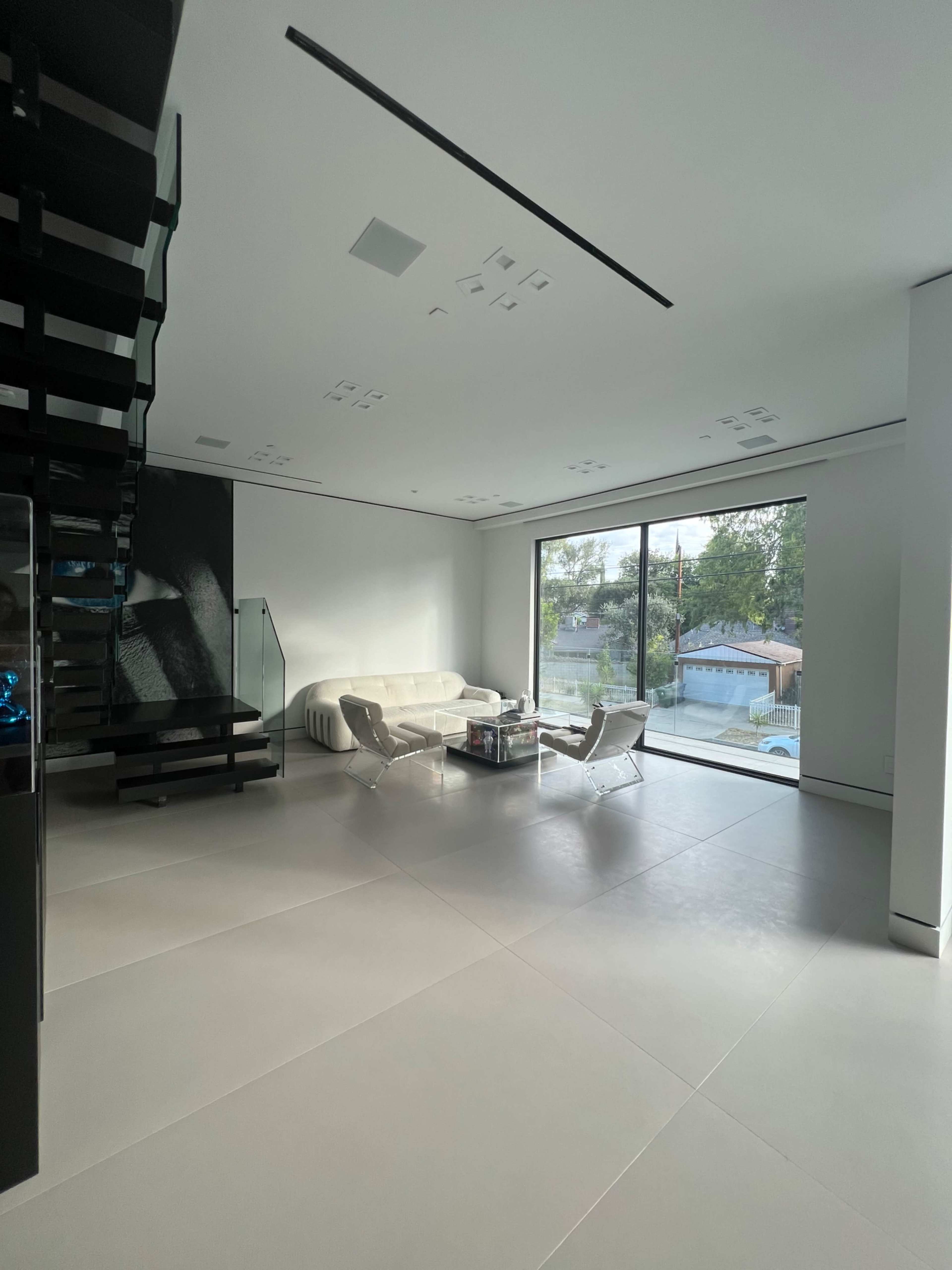 A modern living room with a minimalist design, featuring a white sofa, two sleek chairs, a coffee table, and large windows overlooking a garden.