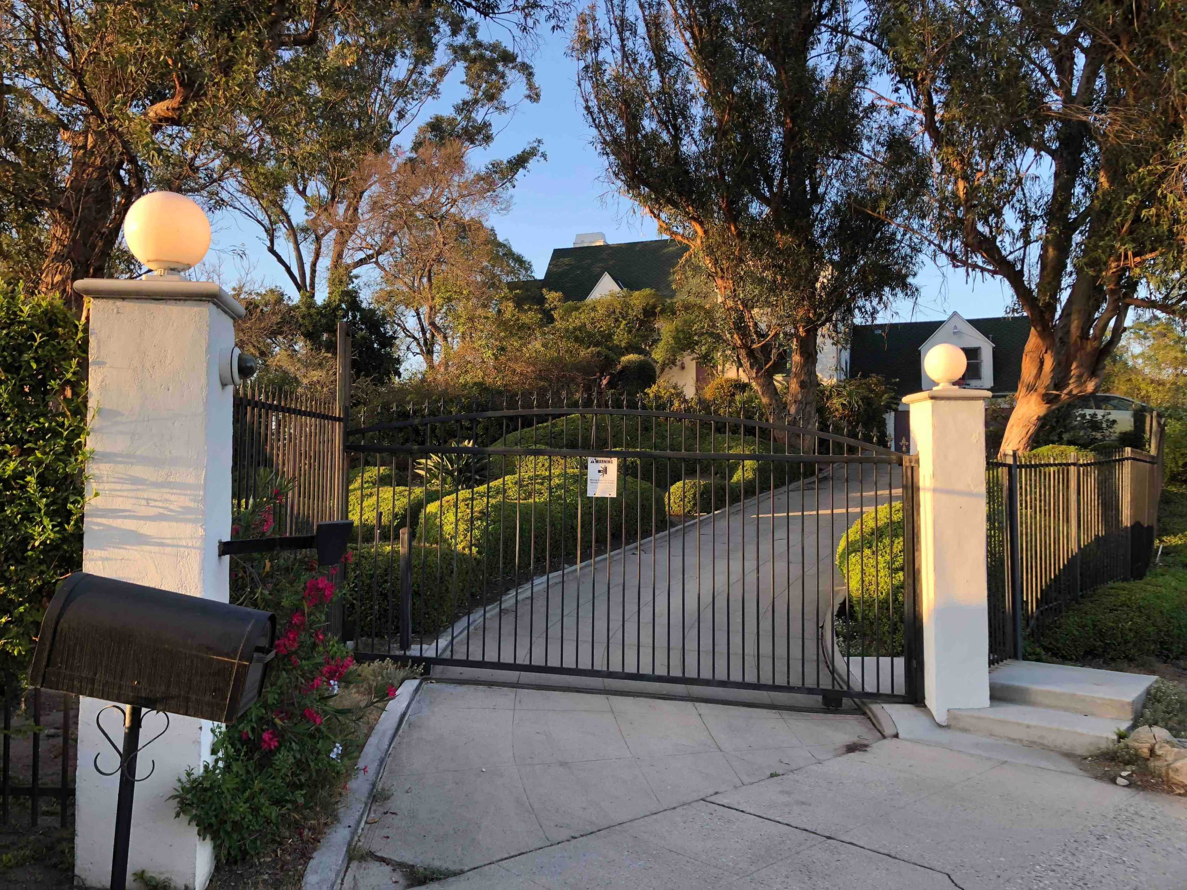 A black wrought iron gate leads up a curved driveway to a house surrounded by landscaped greenery and trees.