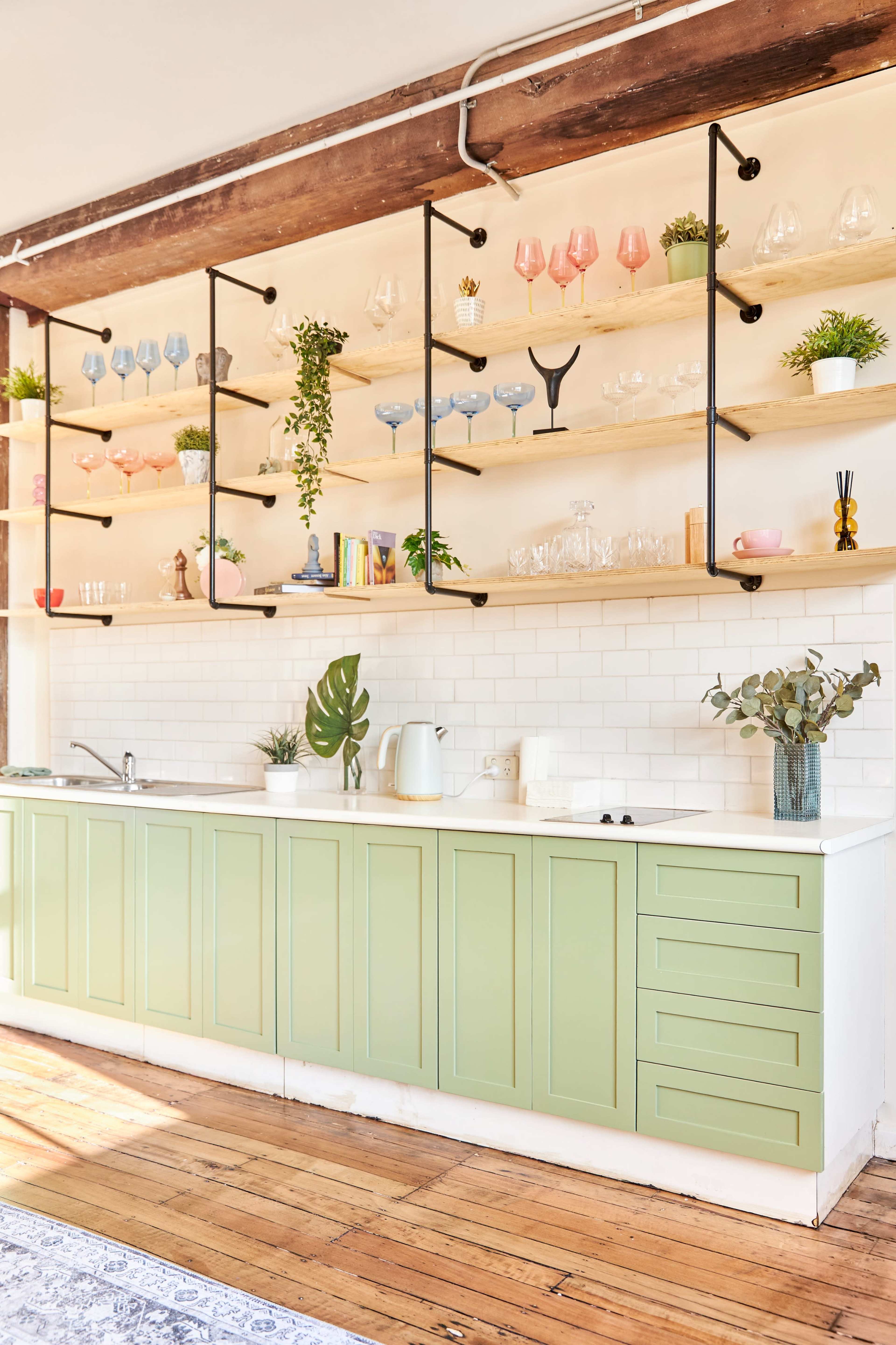 A modern kitchen with light green cabinetry, open shelving displaying various glassware and plants, and a white tiled backsplash.