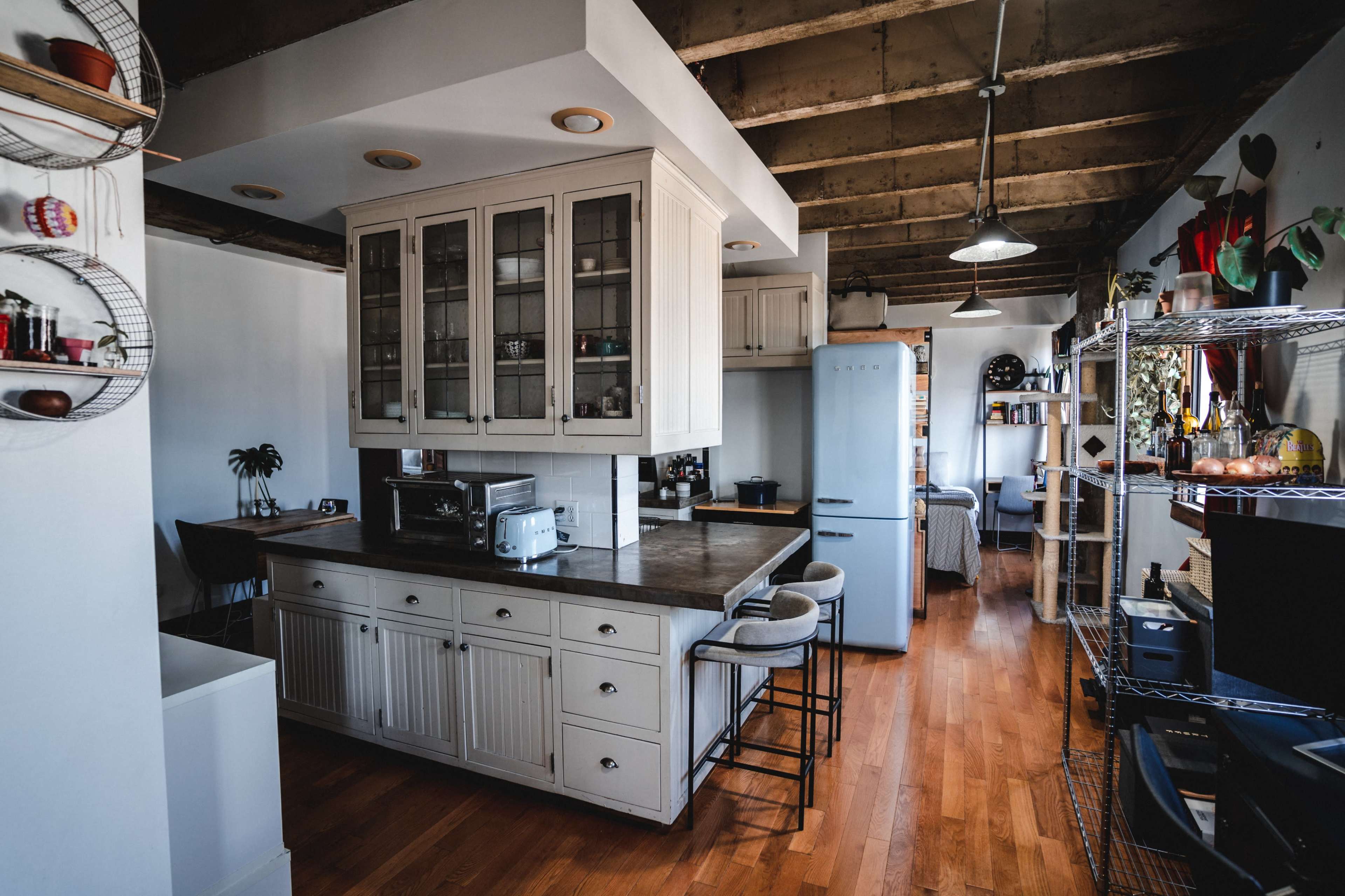 The image shows a streamlined kitchen with white cabinetry, a central island with bar stools, and a refrigerator, all set in a modern space with exposed wooden beams.