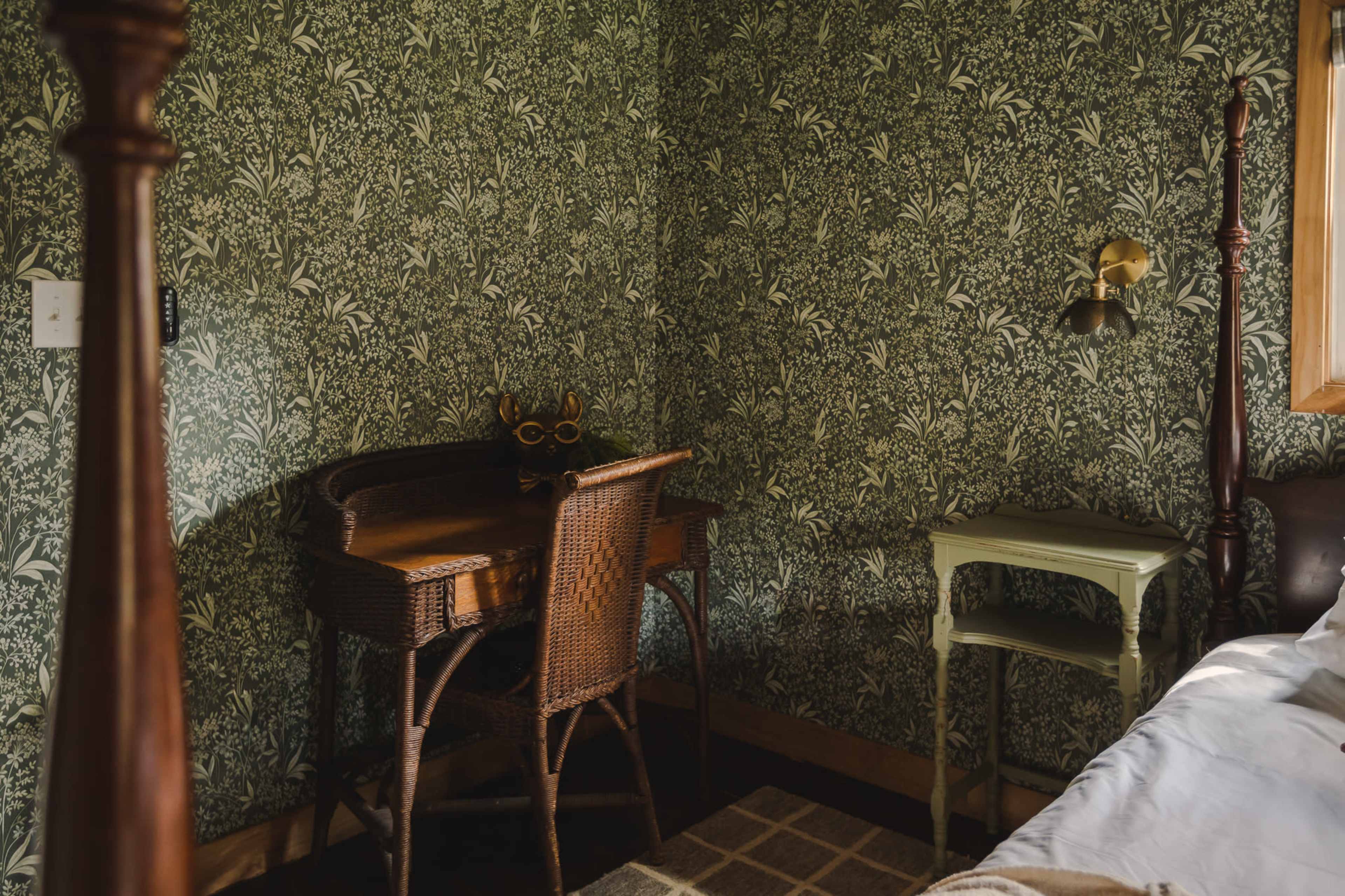 The image shows a corner of a room with patterned green wallpaper, a wooden desk with a wicker chair, and a small light fixture on the wall.