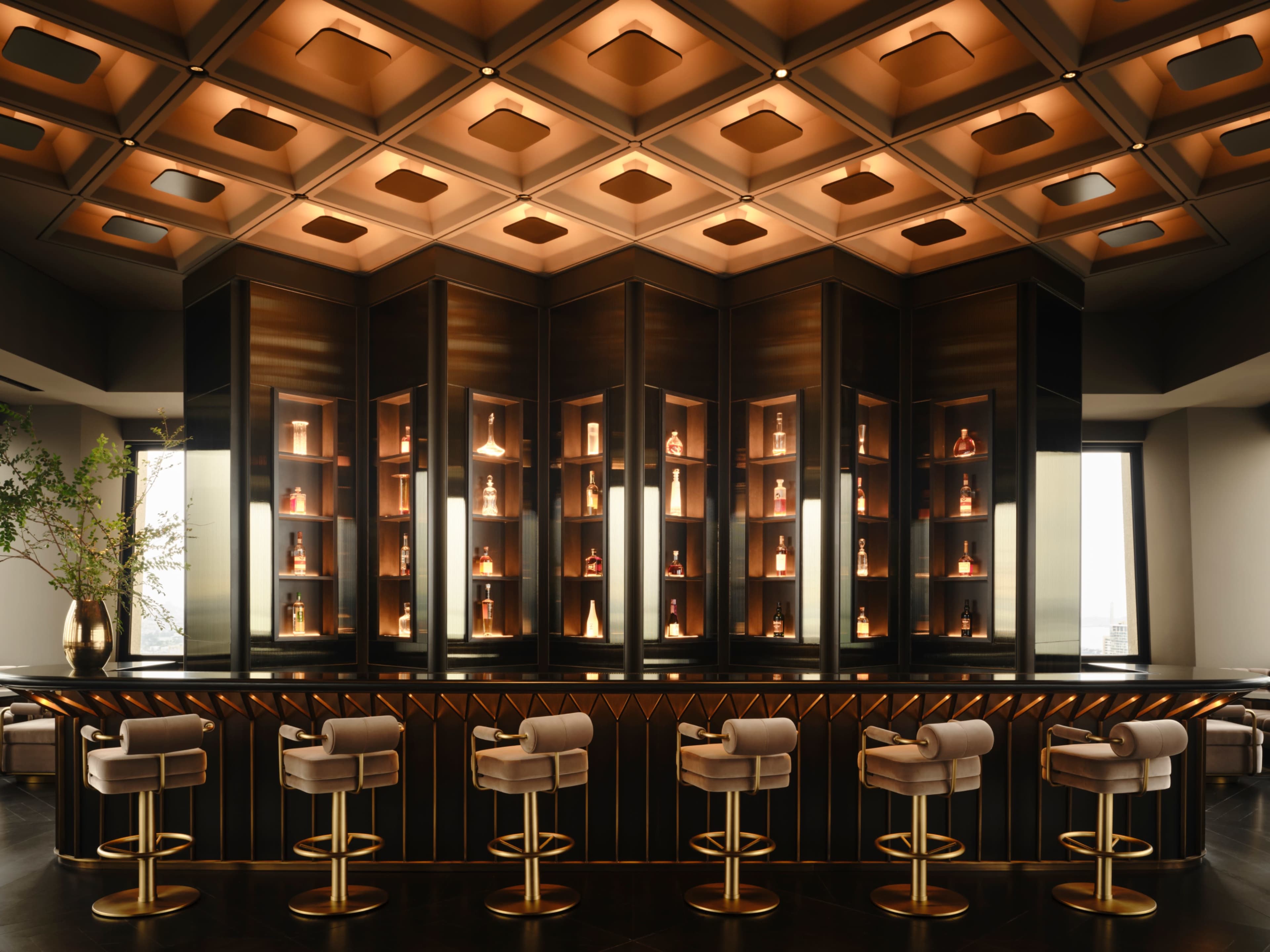 The image shows a modern bar with a geometric design, featuring backlit shelves displaying bottles and a row of upholstered bar stools.