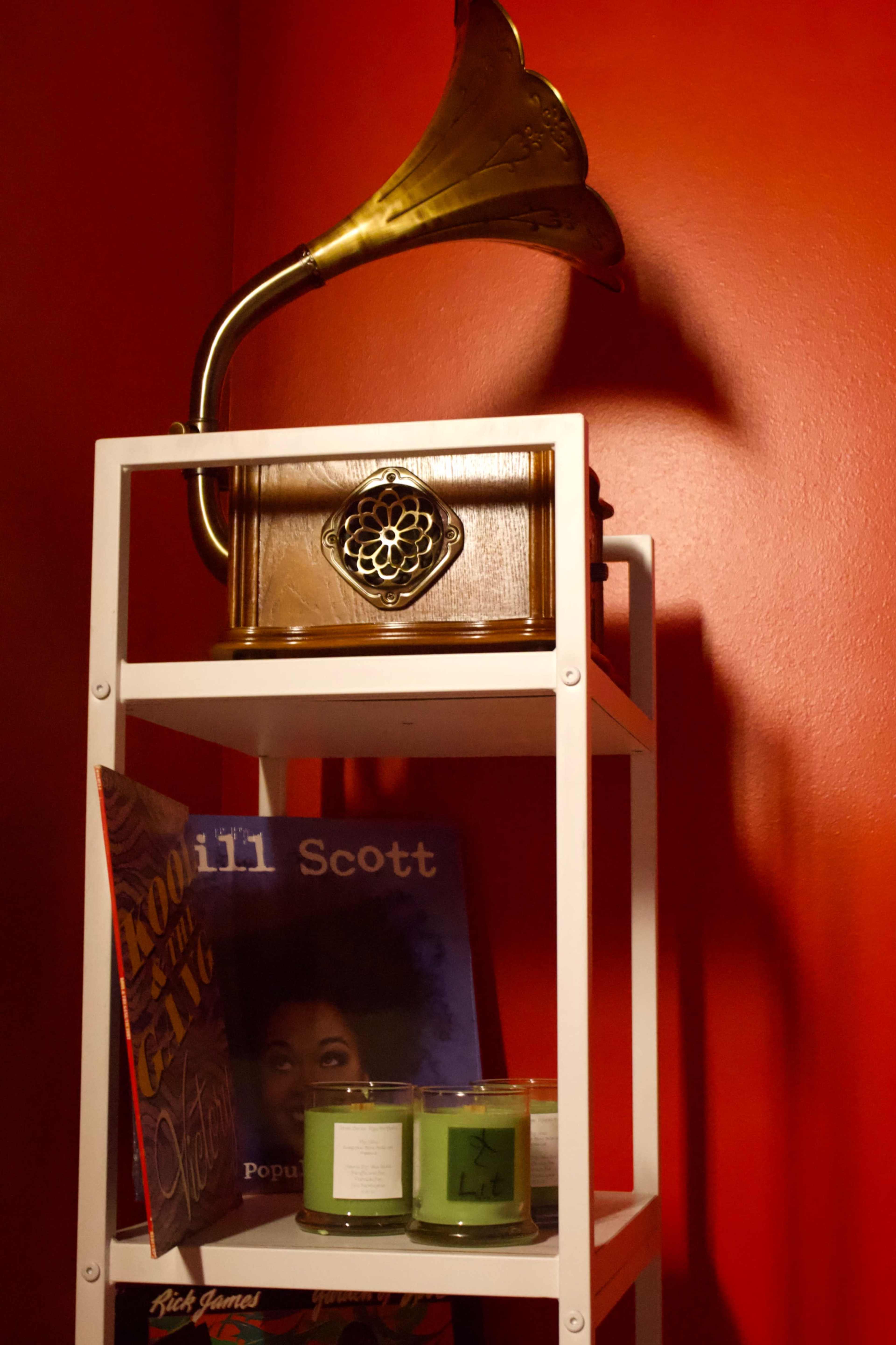 A wooden gramophone sits on a white shelf beside a record by Jill Scott and two green candles against a red wall.
