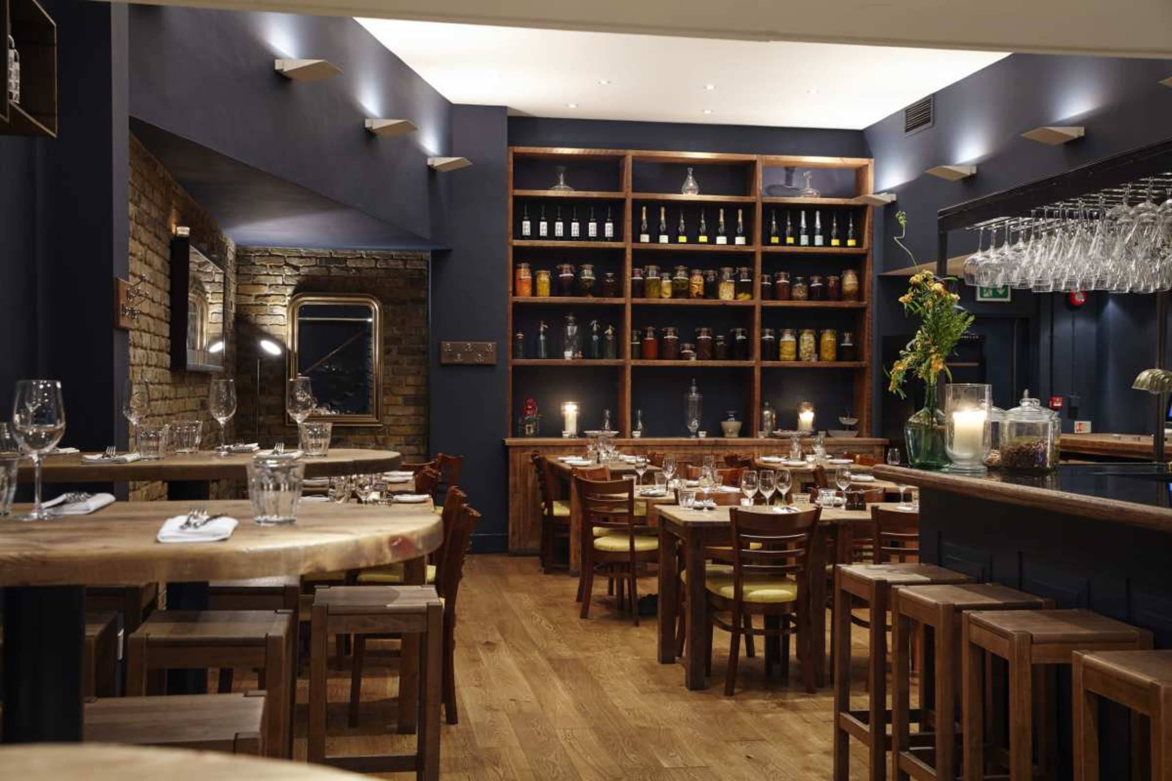 The image depicts a cozy restaurant interior with wooden tables and chairs, shelves lined with jars, and ambient lighting.