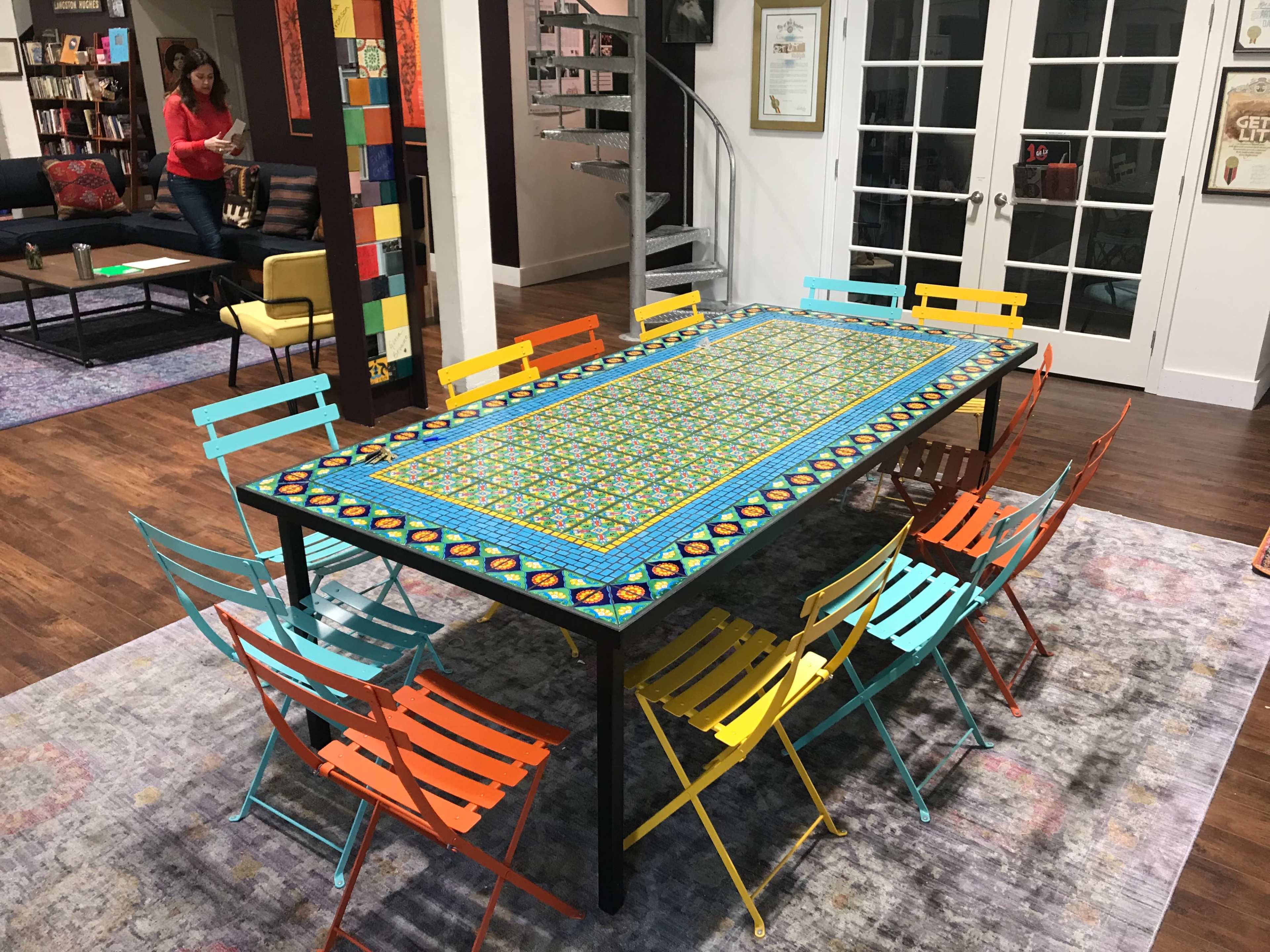 A large table with a colorful patterned tablecloth is surrounded by brightly colored metal chairs in a room featuring spiral stairs and bookshelves.