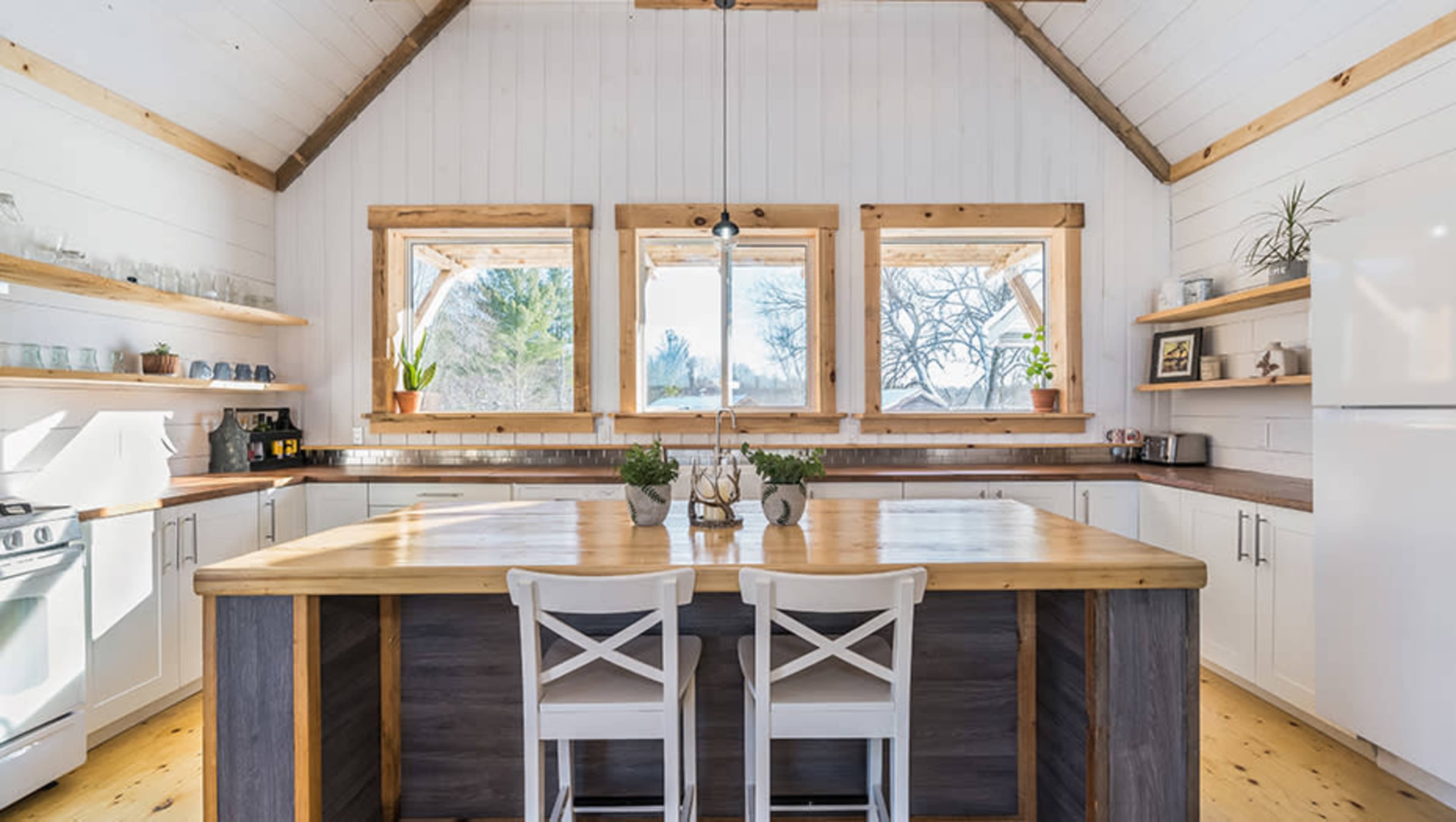 A modern kitchen features a central wooden island with two white chairs, surrounded by white cabinets and large windows that let in natural light.