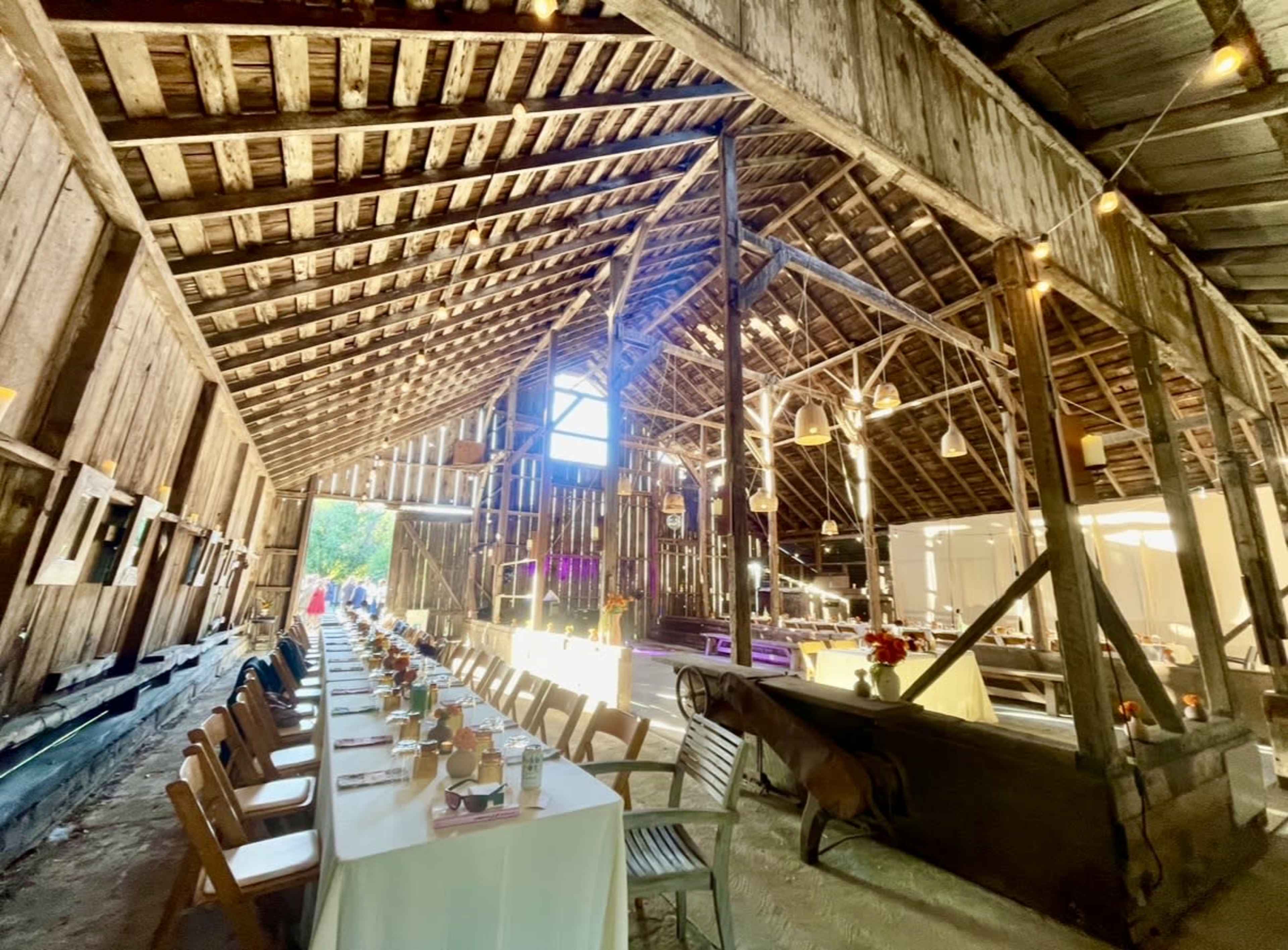 The image shows a long banquet table set with dishes and glasses inside a rustic barn illuminated by hanging lights.