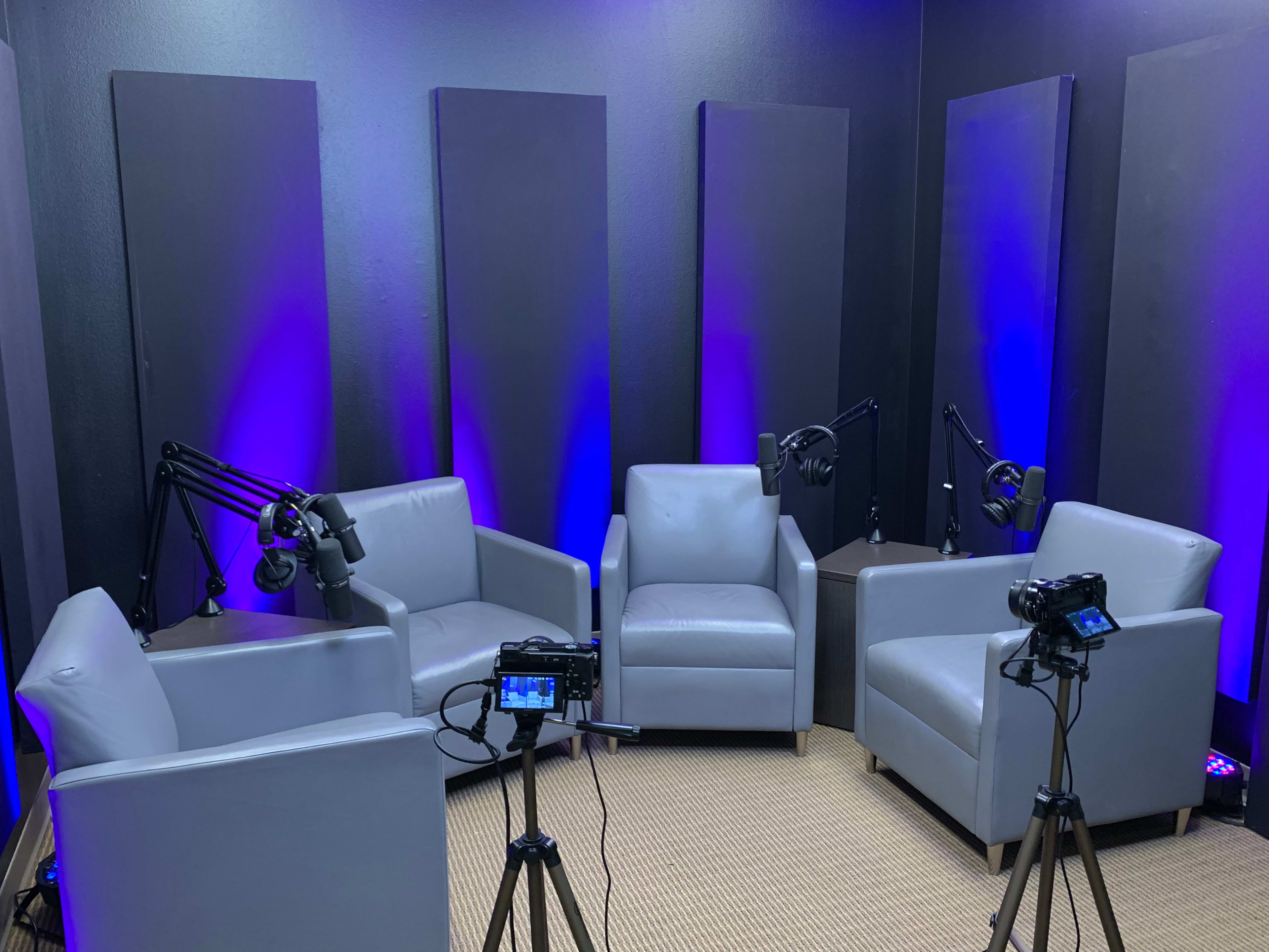 A podcast studio with four gray chairs, two microphones on adjustable arms, and two cameras set up on tripods.