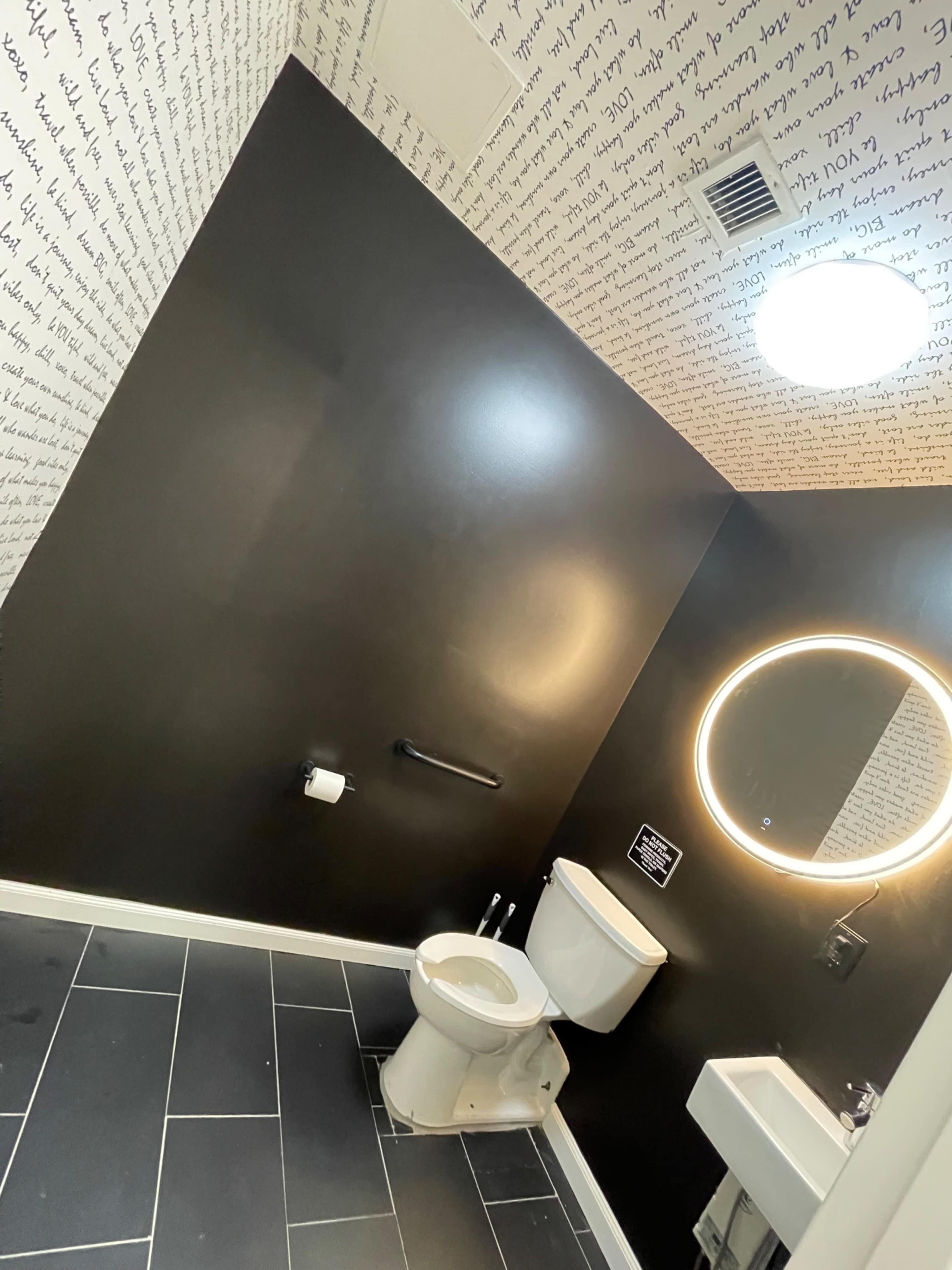 A modern restroom with black walls containing handwritten text, a round mirror, a toilet, and a tiled floor.