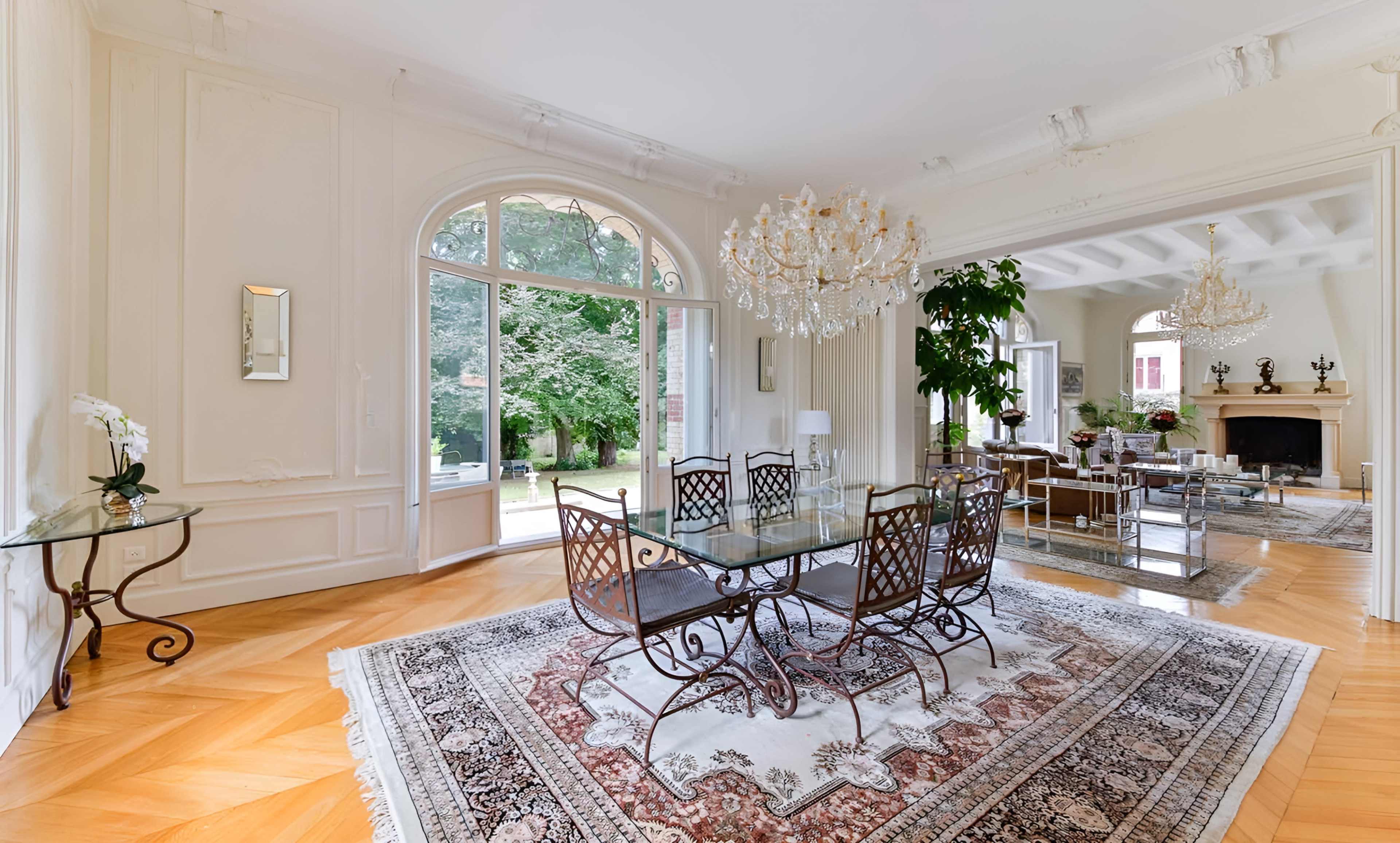 The image shows a spacious dining area with a glass table and metal chairs, surrounded by large windows and elegant decor, leading to a well-maintained garden.