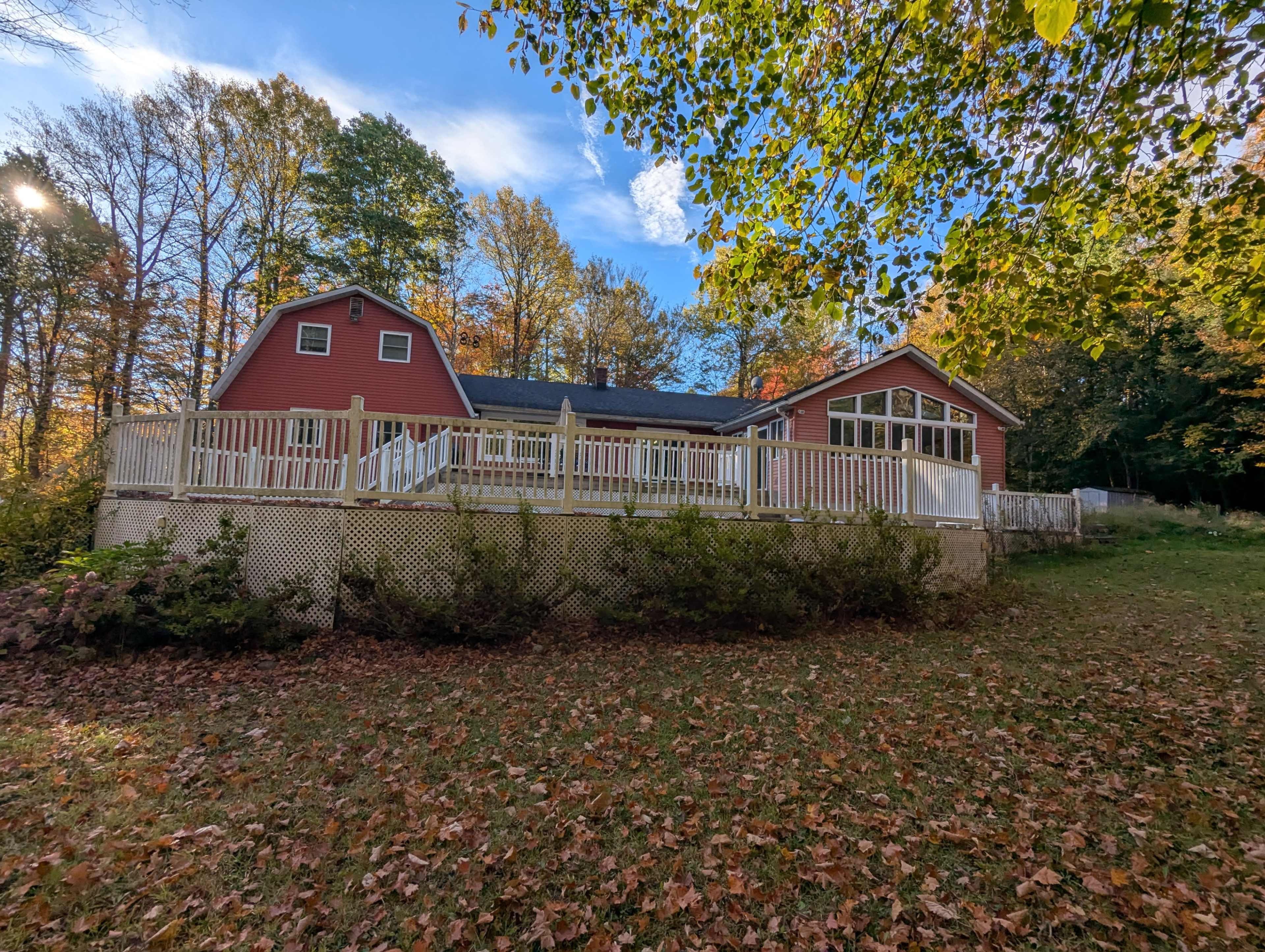 A red house with a white-railed deck is surrounded by trees with autumn leaves on the ground.