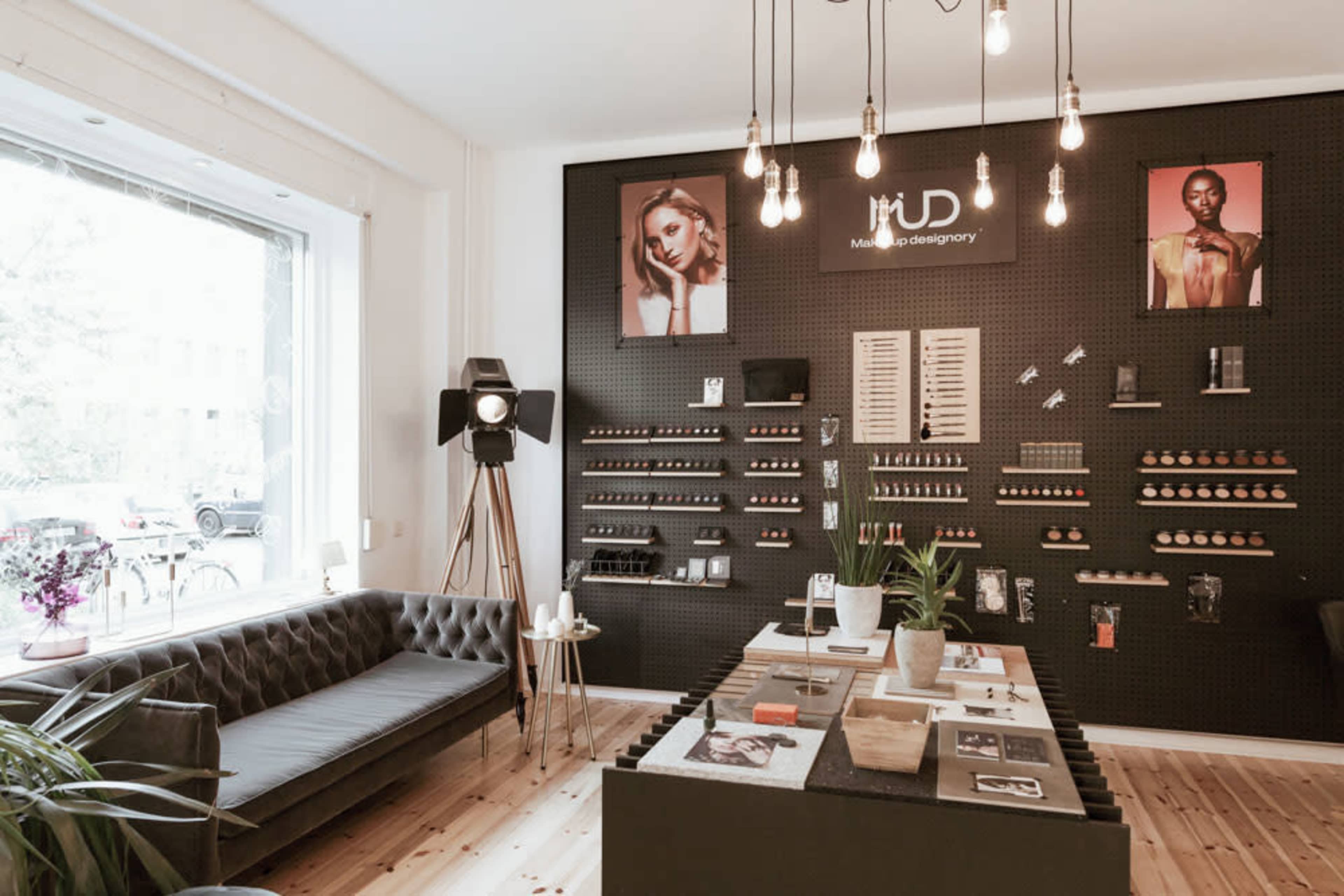 A modern beauty studio features a dark pegboard wall displaying makeup products, a central table with various items, and a cozy seating area.