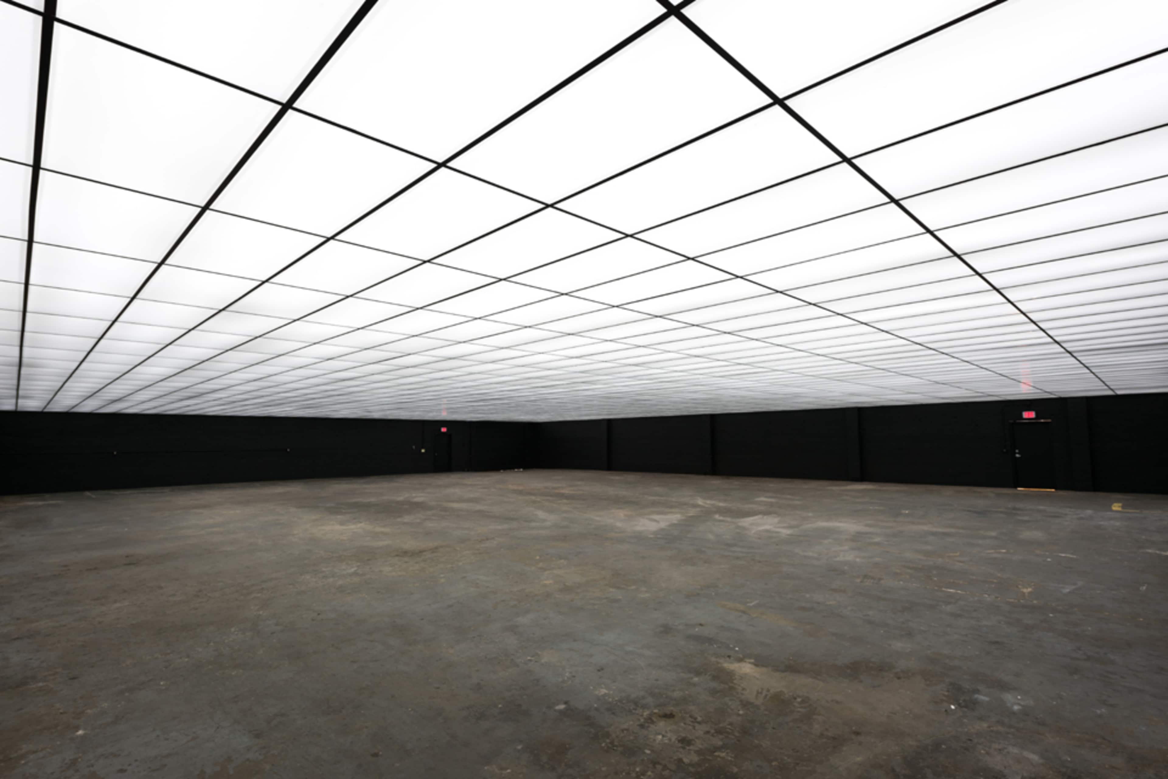 A large, empty room with a flat, illuminated ceiling and dark walls.