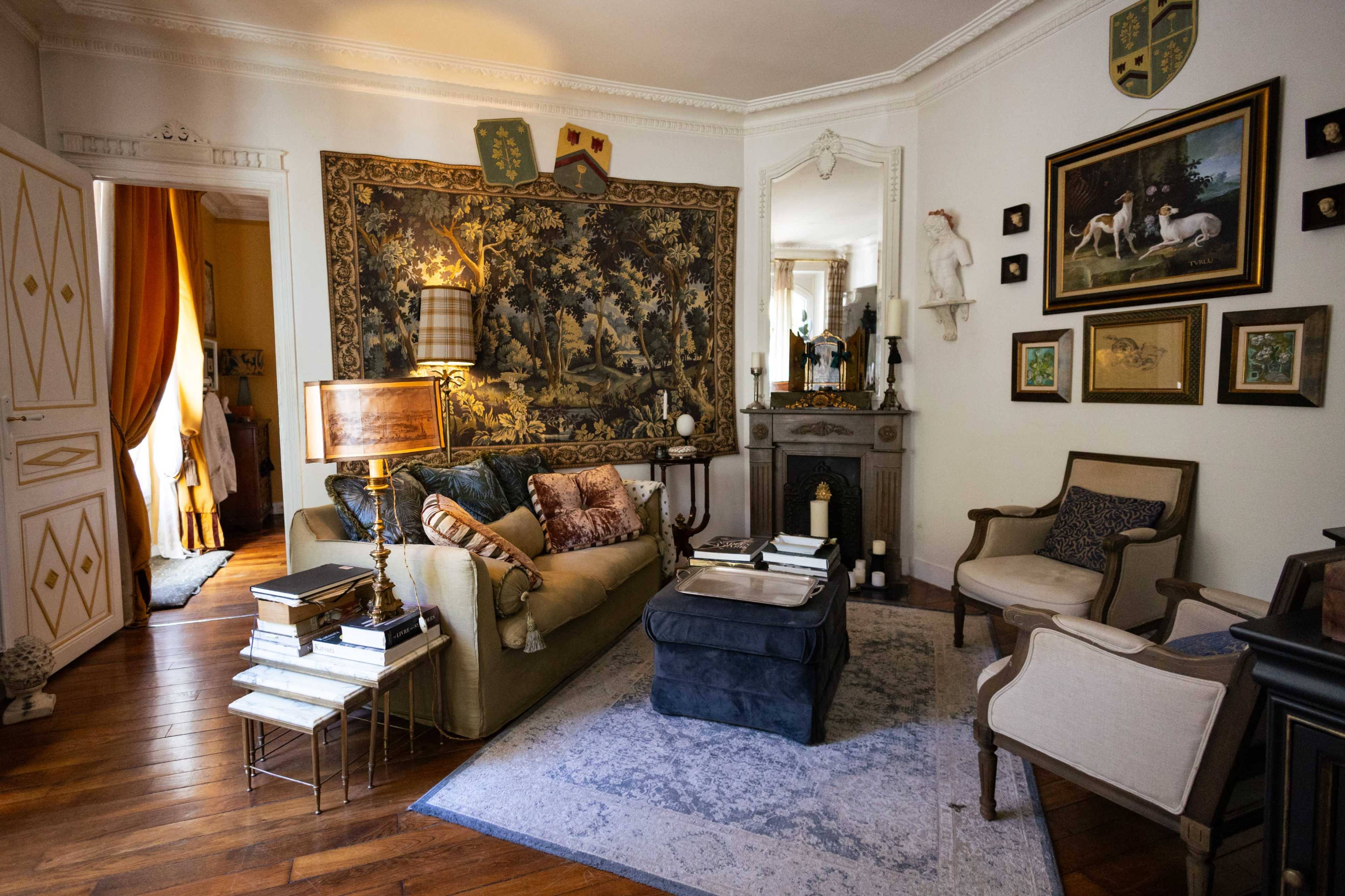 The image shows a richly decorated living room featuring a tapestry on the wall, a blend of vintage and modern furniture, and a coffee table surrounded by seating.