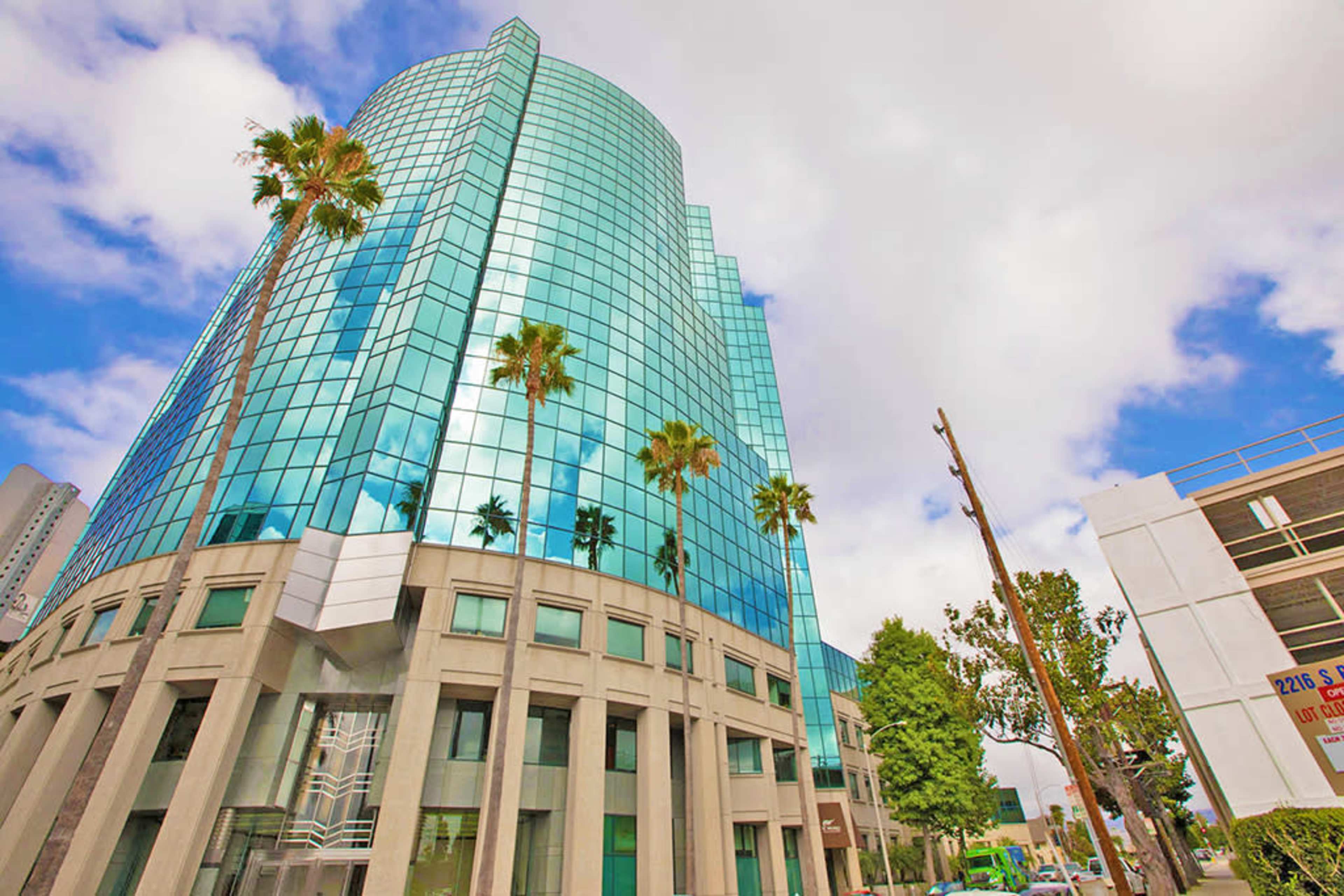 A modern glass building with palm trees in the foreground is set against a partly cloudy sky.