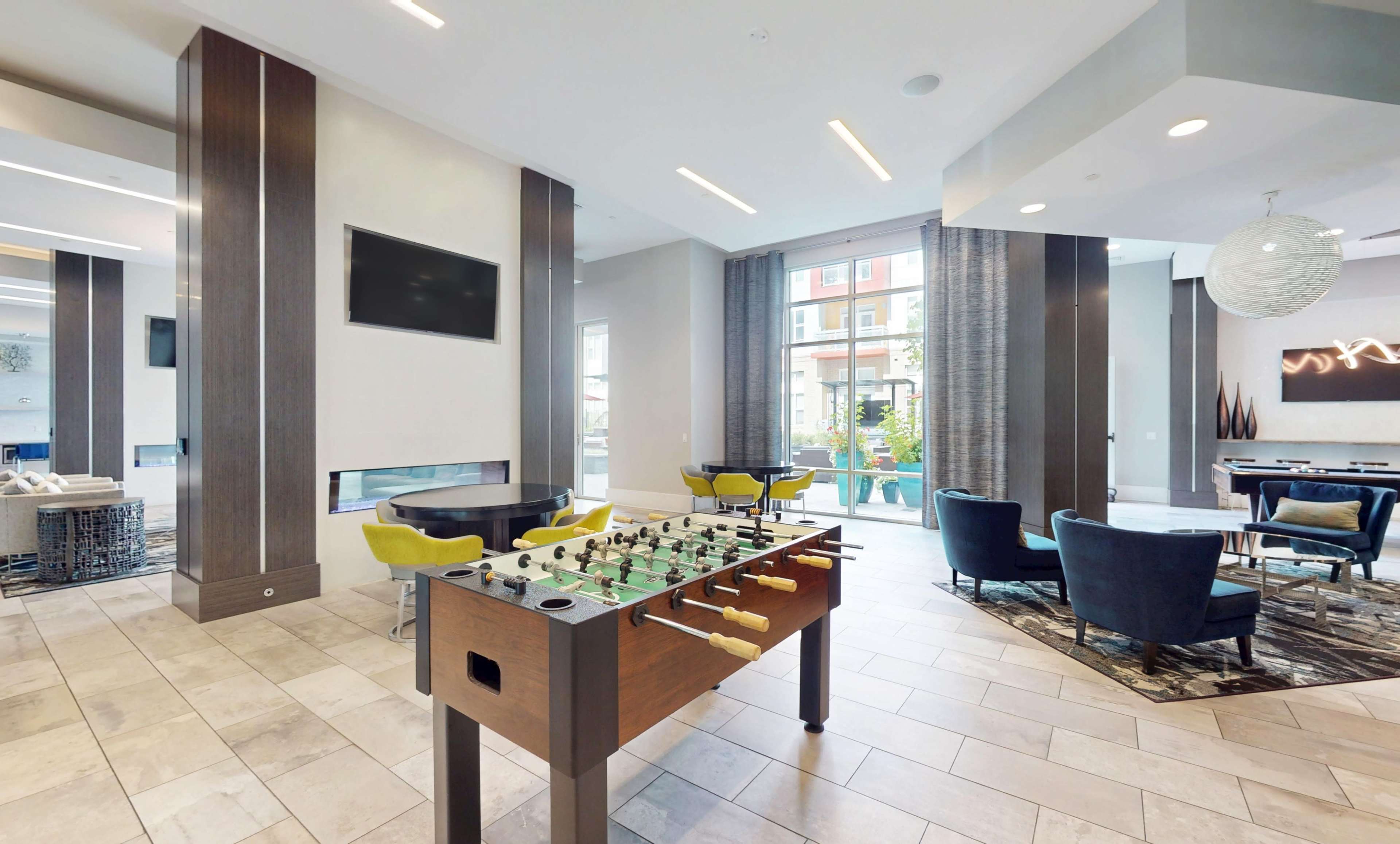 The image shows a lounge area featuring a foosball table, seating, a television, and large windows that provide natural light.