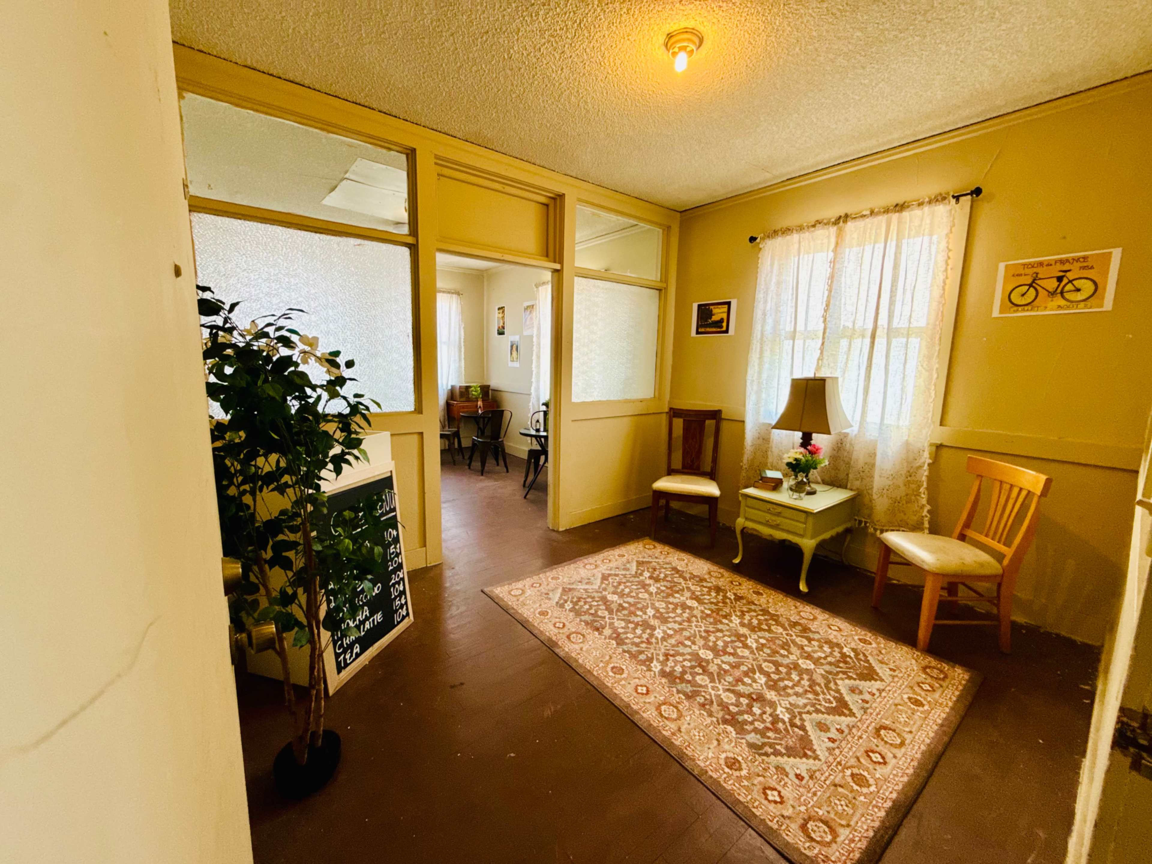 The image shows a well-lit hallway with a patterned rug, potted plant, and seating areas leading into an open room with additional furniture visible.
