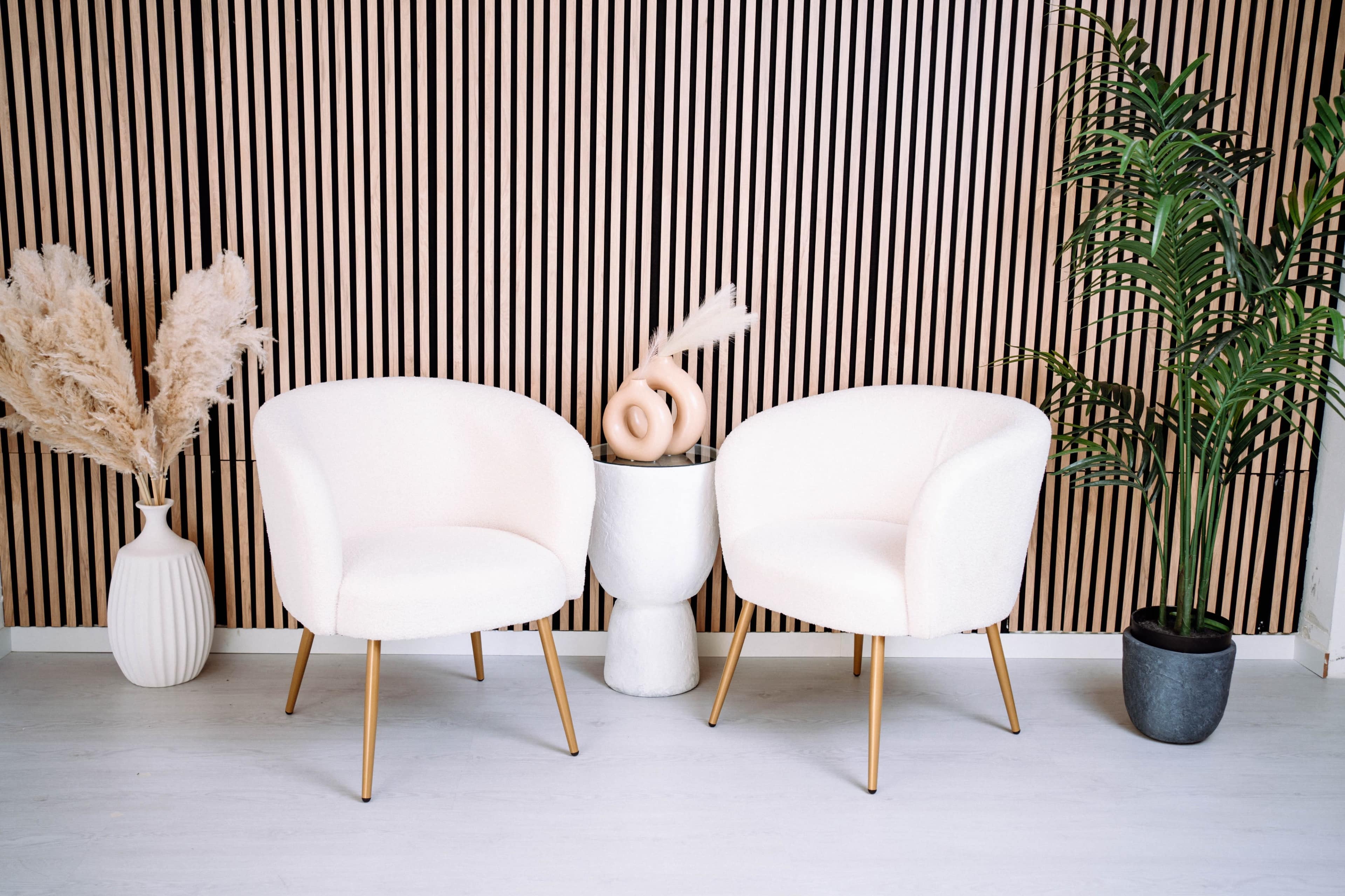The image features two white, upholstered chairs positioned beside a cylindrical table, with decorative elements including a sculptural piece and potted plants against a striped wooden wall.