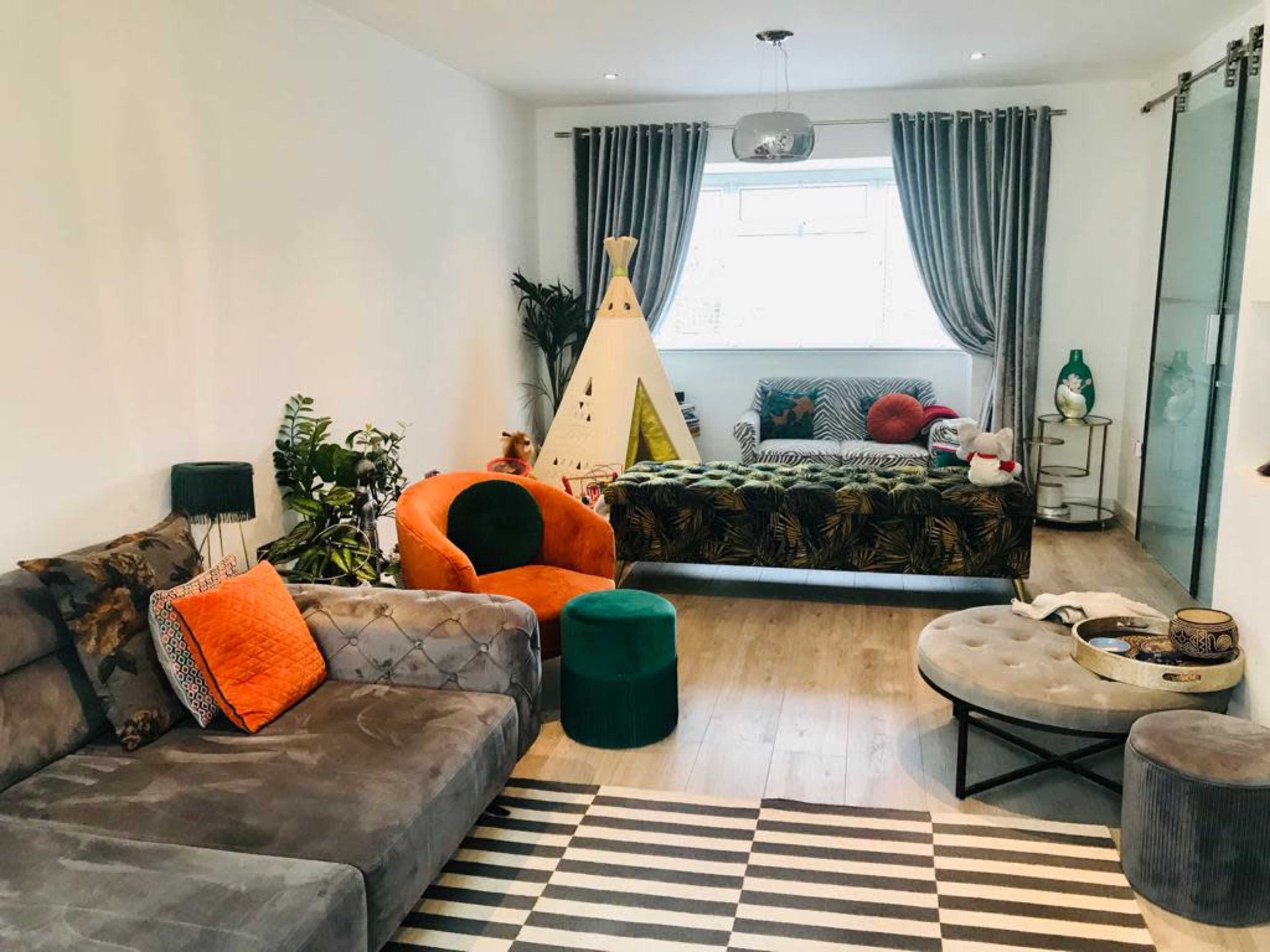 The living room features a mix of seating options, including a gray sofa, an orange chair, and a patterned couch, with a children's teepee and various plants accentuating the space.
