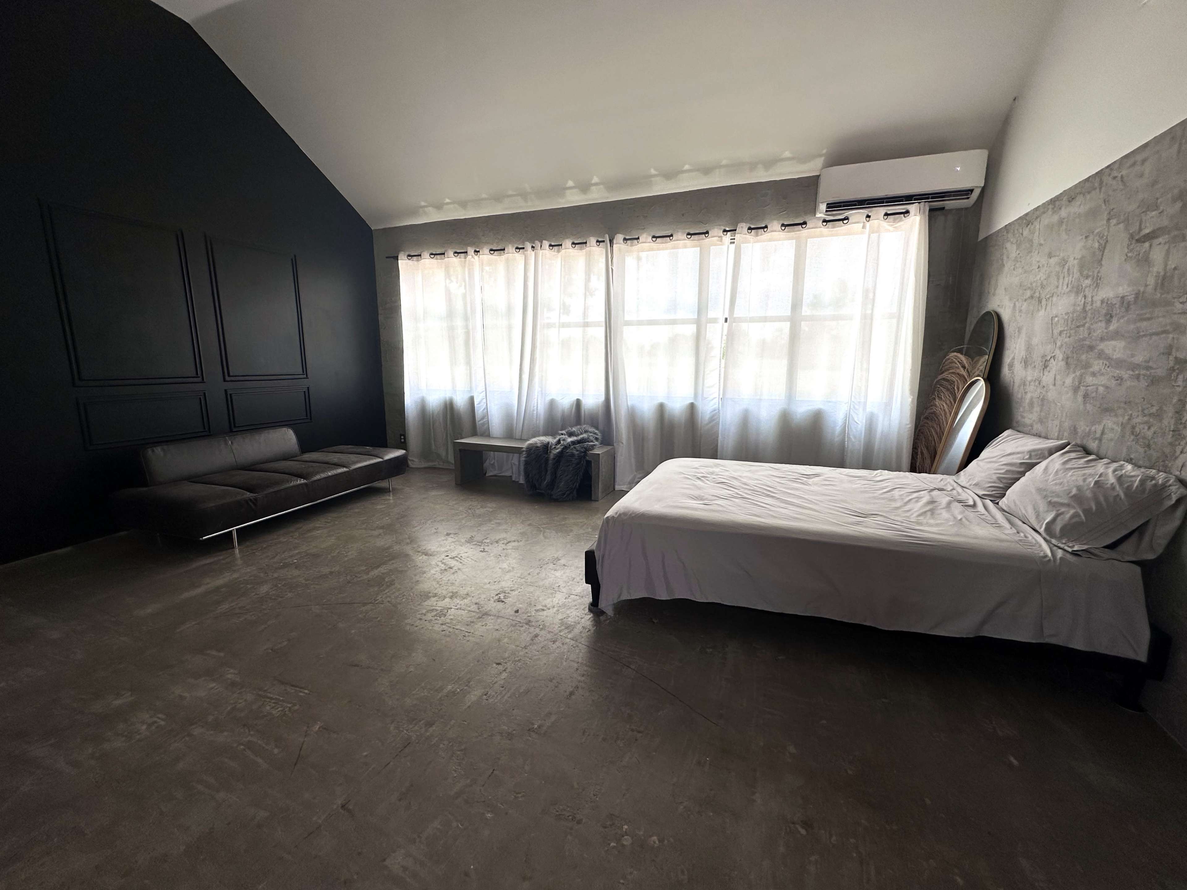A modern bedroom features a bed, a sleek sofa, a coffee table, and large windows covered with sheer curtains, all set against dark and light walls.