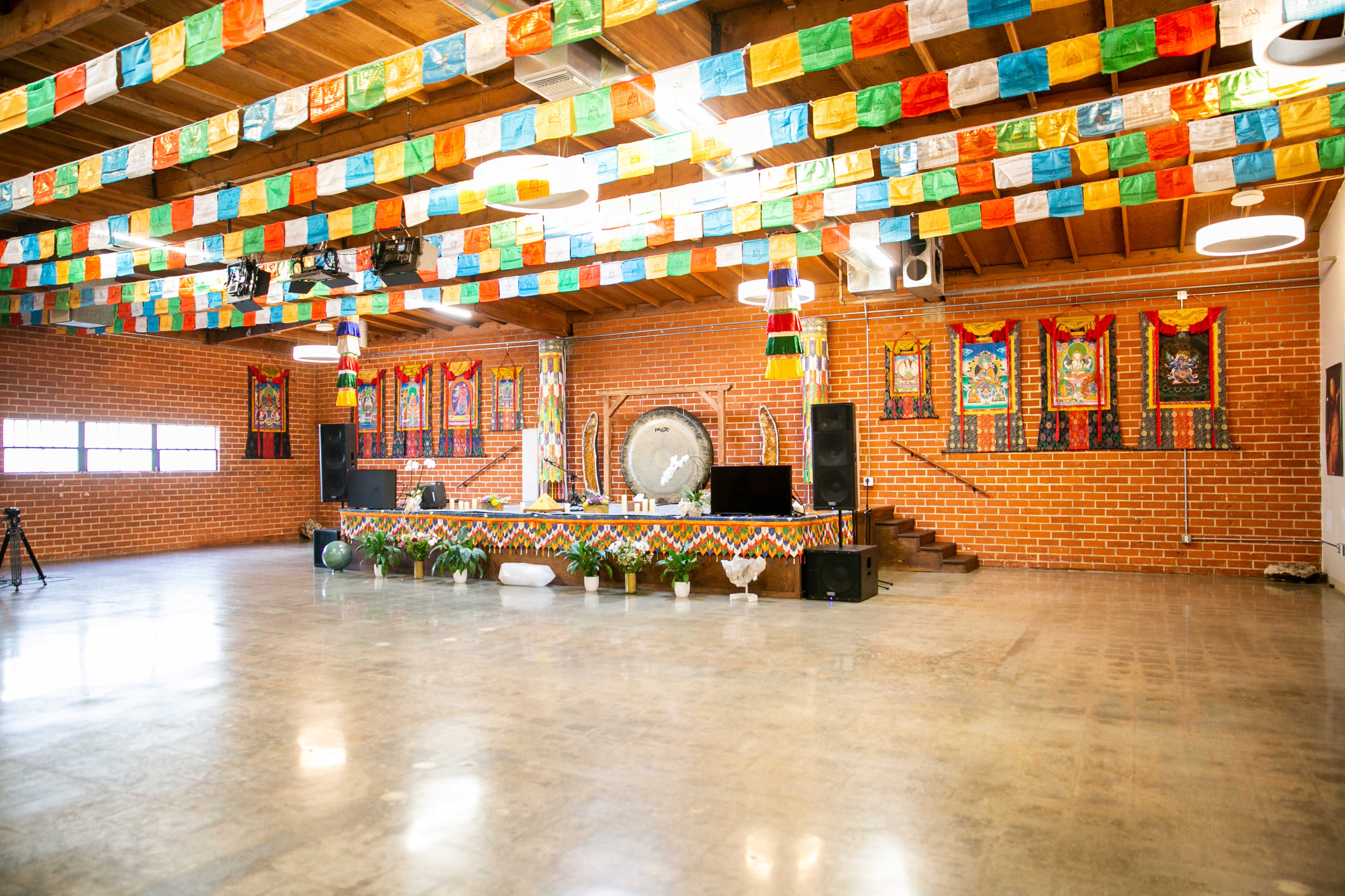 A spacious indoor venue with a stage decorated with colorful banners, lush plants, and a large gong, all set against brick walls and wooden beams.