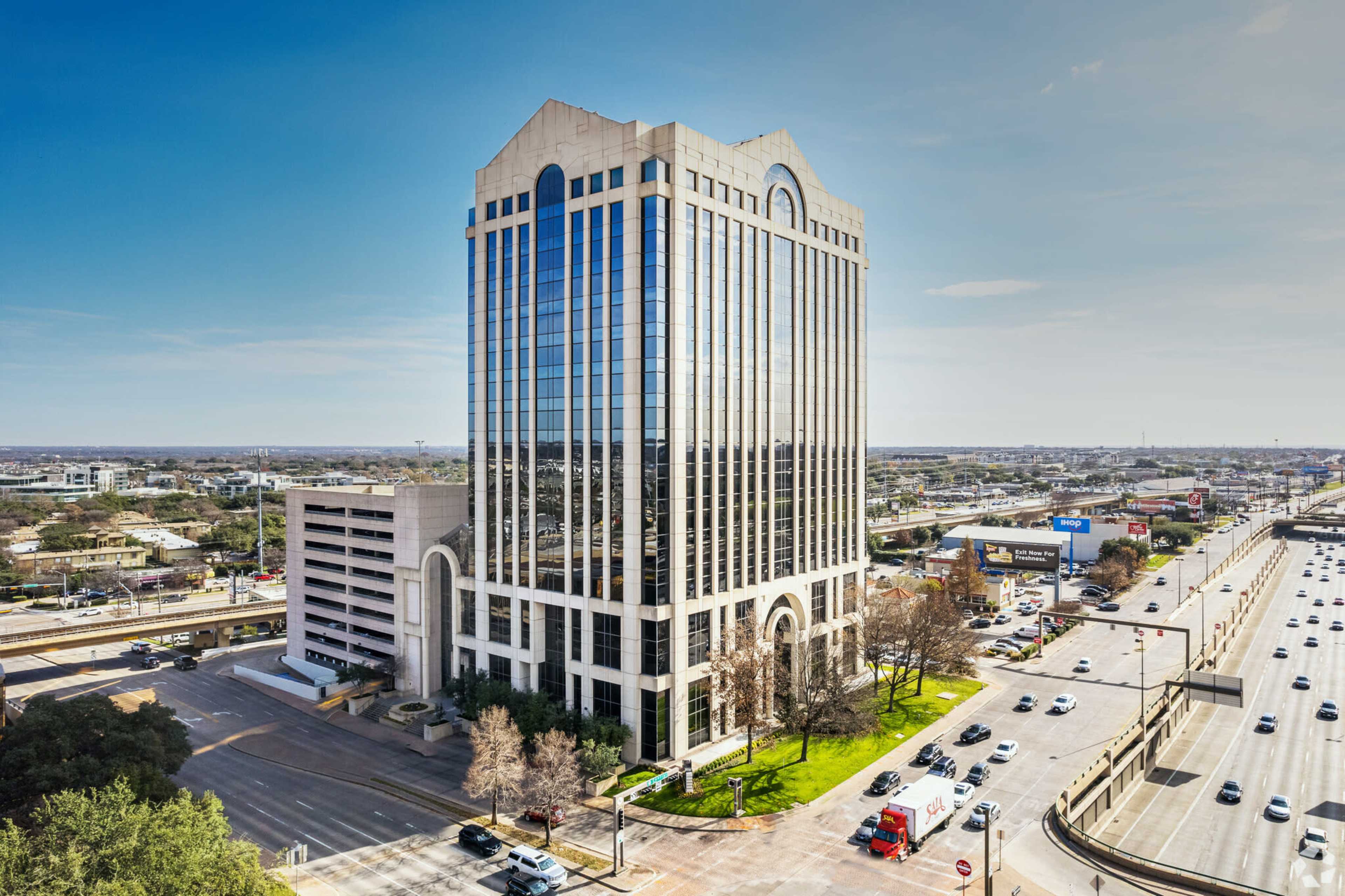 A tall glass office building stands alongside a busy highway, with multiple vehicles on the road and a commercial area in the background.